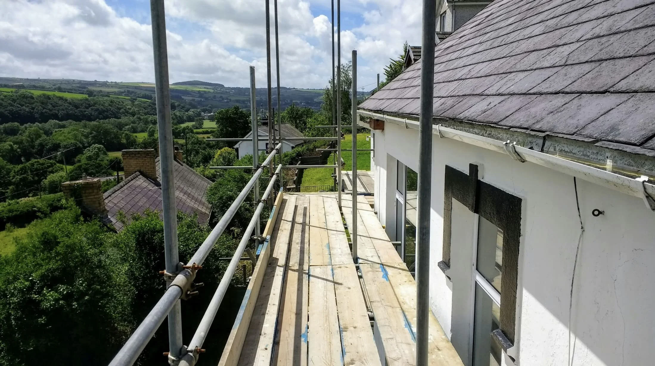 Scaffolding on the side of a white building under construction with a view of a rural landscape with green fields and hills in the background.