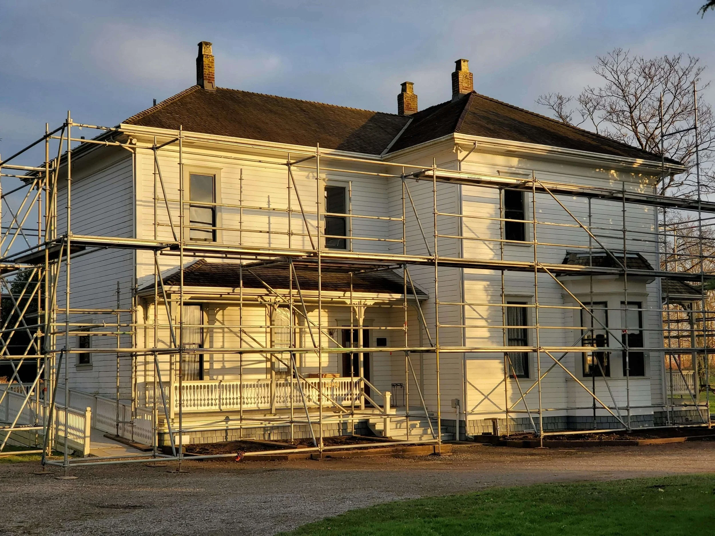 A two-story white house undergoing renovation with metal scaffolding surrounding it during sunset.