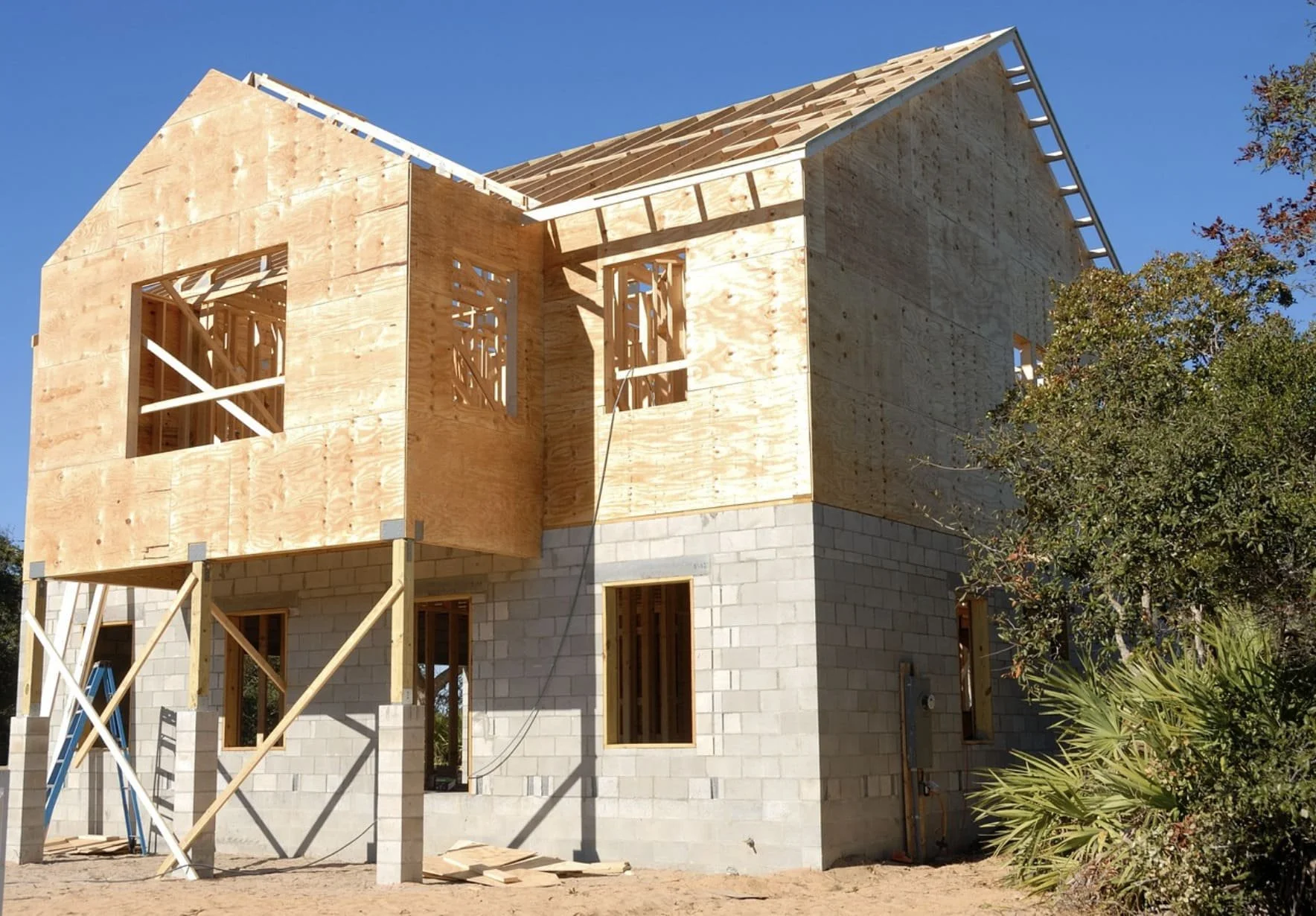 A two-story house under construction with wooden framing and a concrete block foundation, with a clear blue sky and trees nearby.