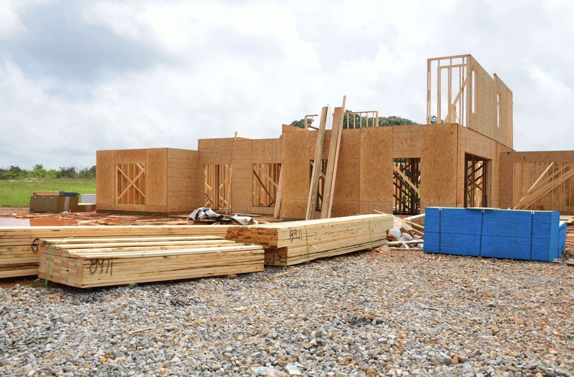 Construction site with wooden framing for a house, piles of lumber, and building materials on a gravel ground, under a cloudy sky.