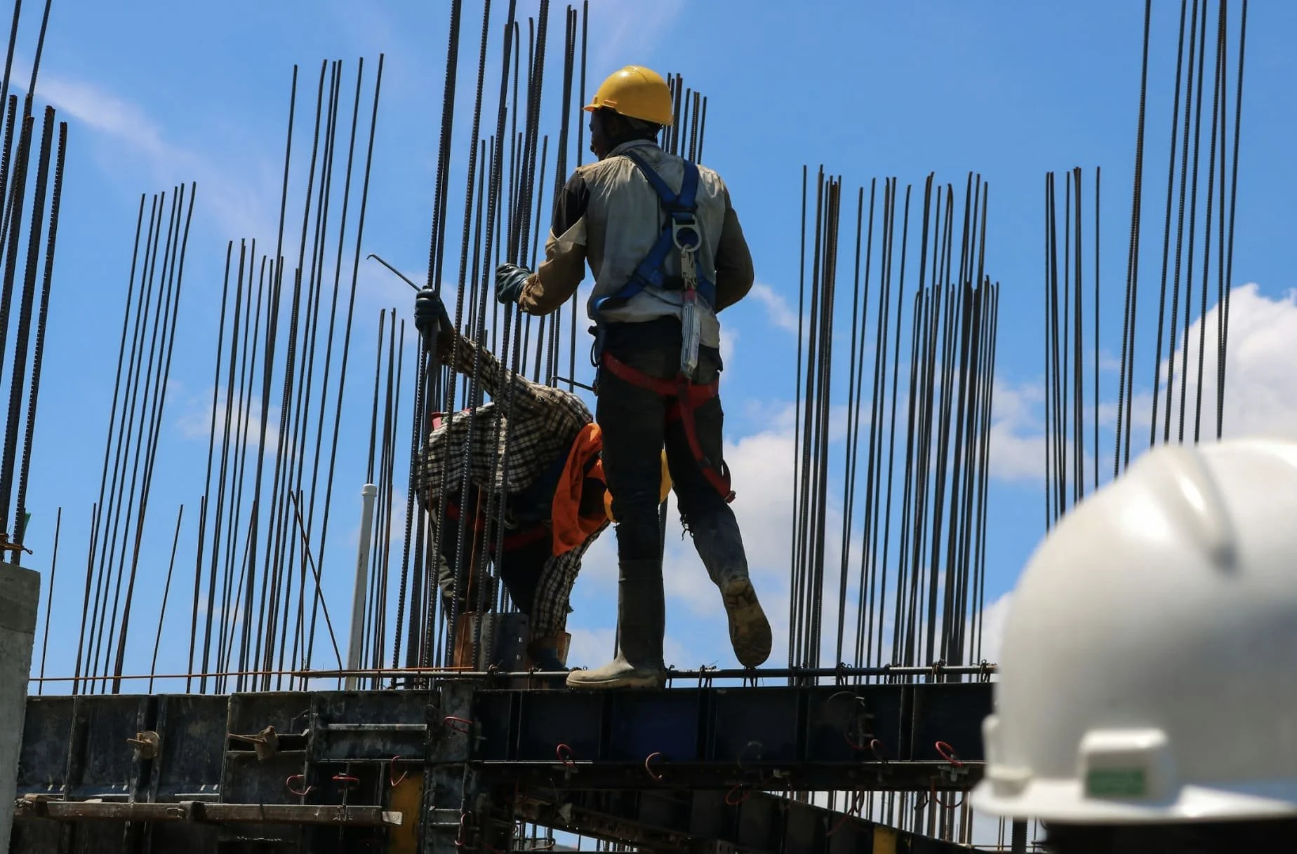 Two construction workers working on a building site, standing on a concrete platform with steel reinforcement bars around them. One worker is wearing a yellow safety helmet, harness, and gloves, holding a tool, while the other is bent over working on the rebar, with a safety vest and checkered shirt. A white safety helmet is visible in the foreground, with a blue sky and clouds in the background.