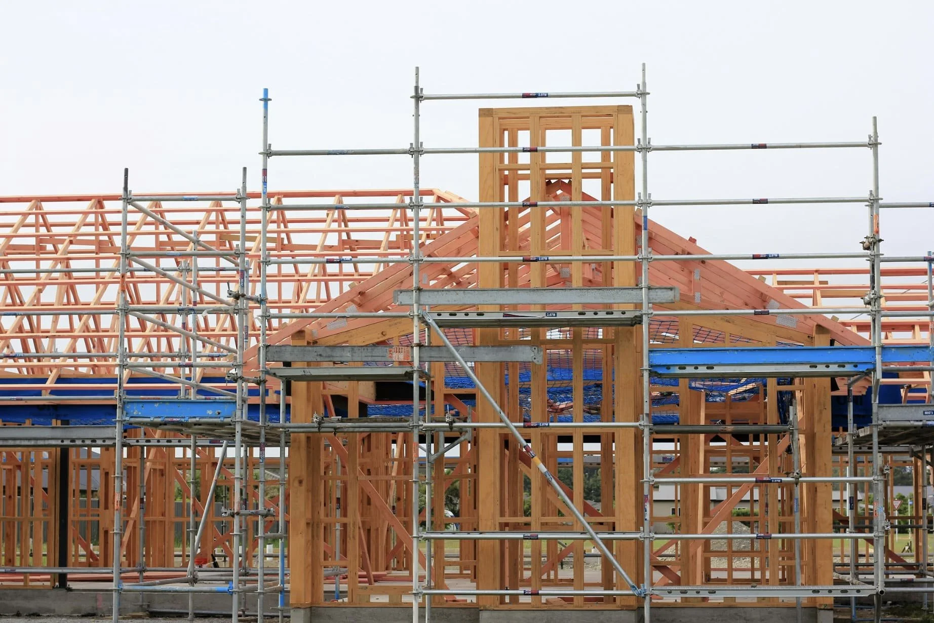 Construction site where a wooden house frame is being built, with scaffolding surrounding it and a cloudy sky in the background.