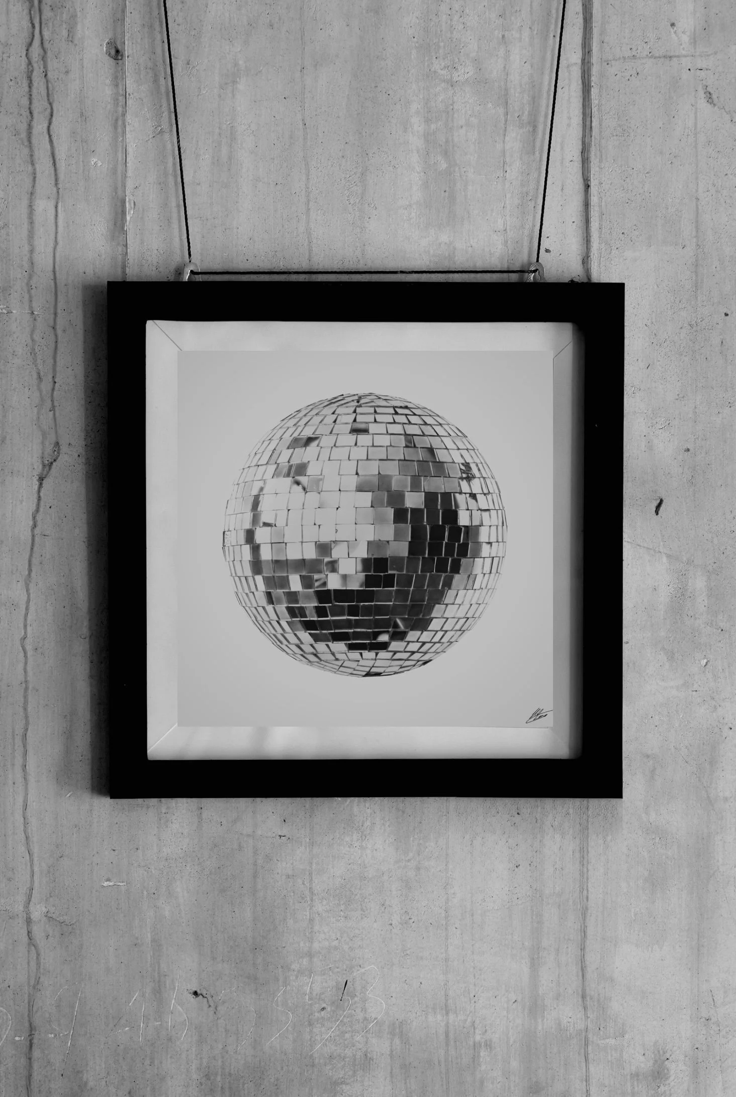 A framed black-and-white photograph of a mirrored disco ball hanging on a black frame, displayed against a concrete wall.
