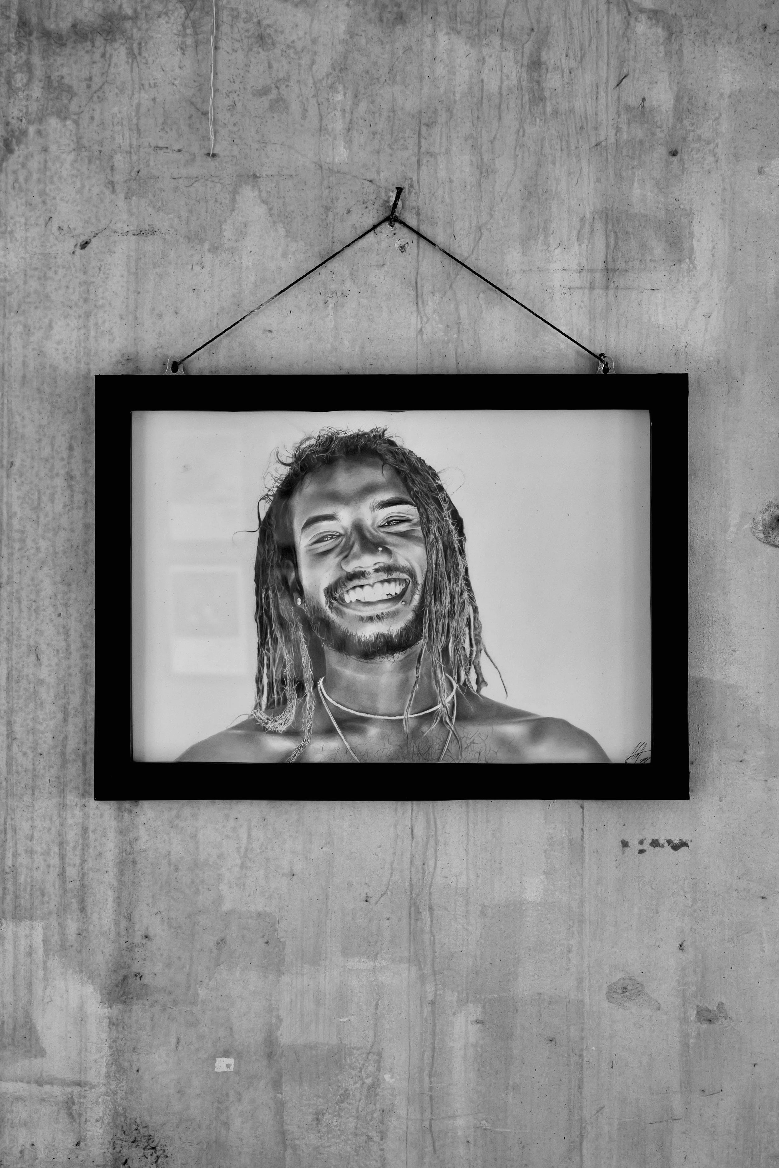 Black-and-white photograph of a framed portrait of a smiling man with dreadlocks, hanging on a rough concrete wall.