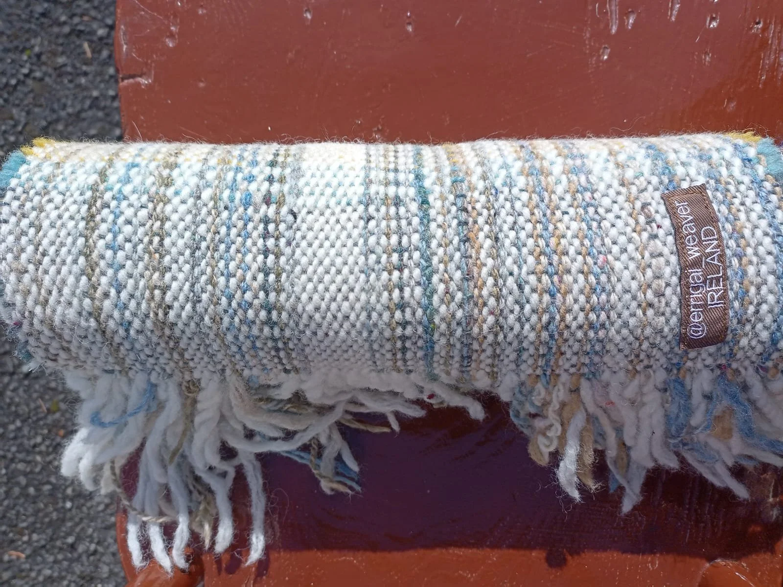 A rolled woven rug with fringes at the ends and a tag that reads '@erigan WEAVE IRELAND' resting on a wooden surface.