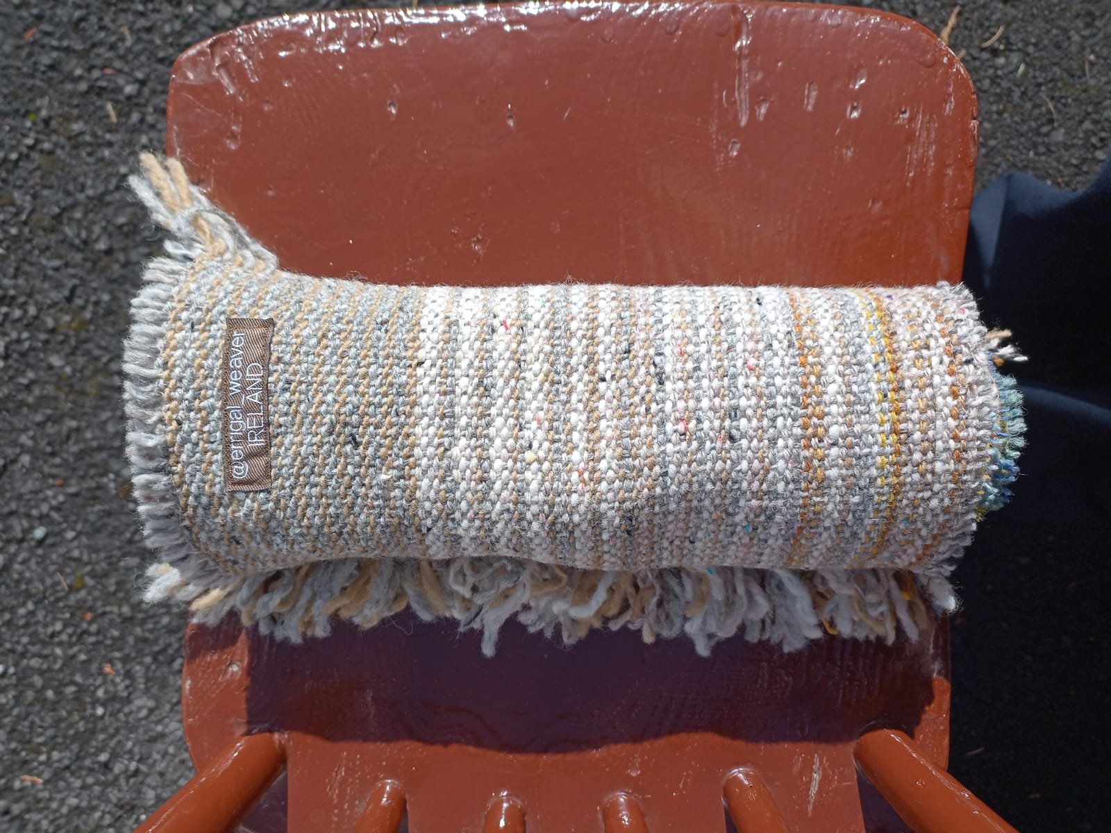 A rolled-up, multicolored woven fabric or towel placed on a brown chair outdoors on gravel ground.