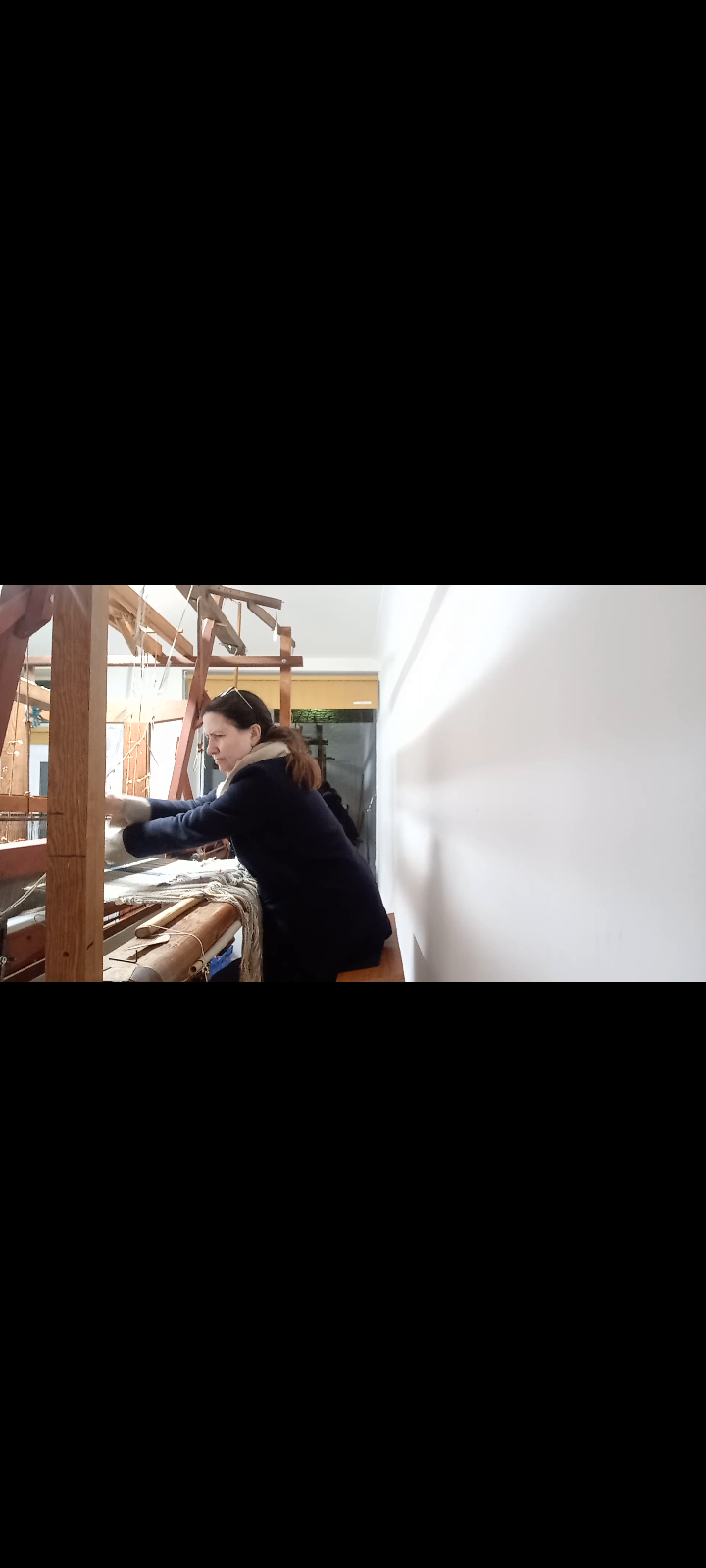 Woman working on a wooden loom, placing yarn or fabric, in a workshop with white walls.