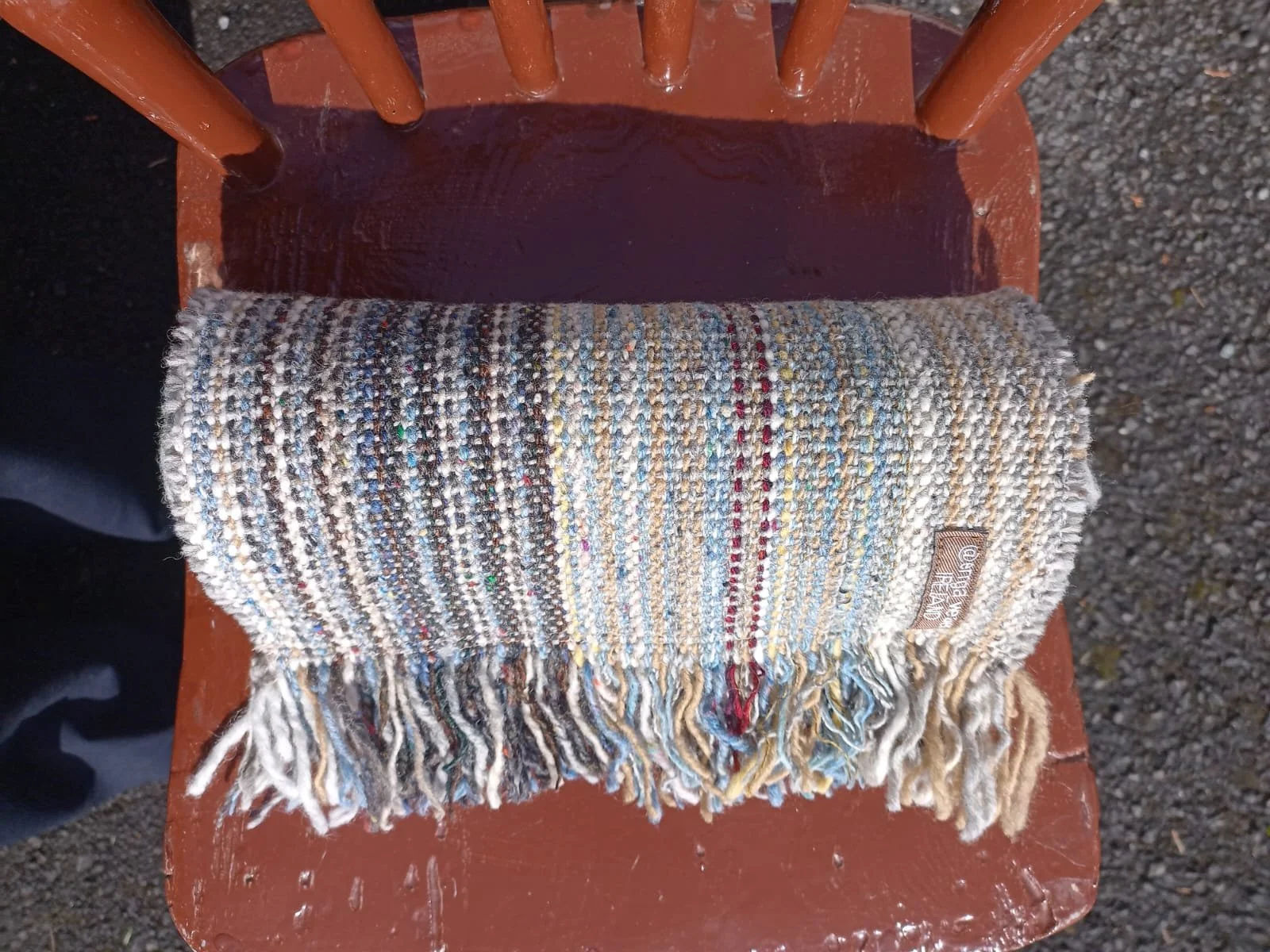 A multicolored striped woven scarf with fringe on the bottom, placed on a brown wooden chair with orange legs. The chair is outdoors on a gravel surface.