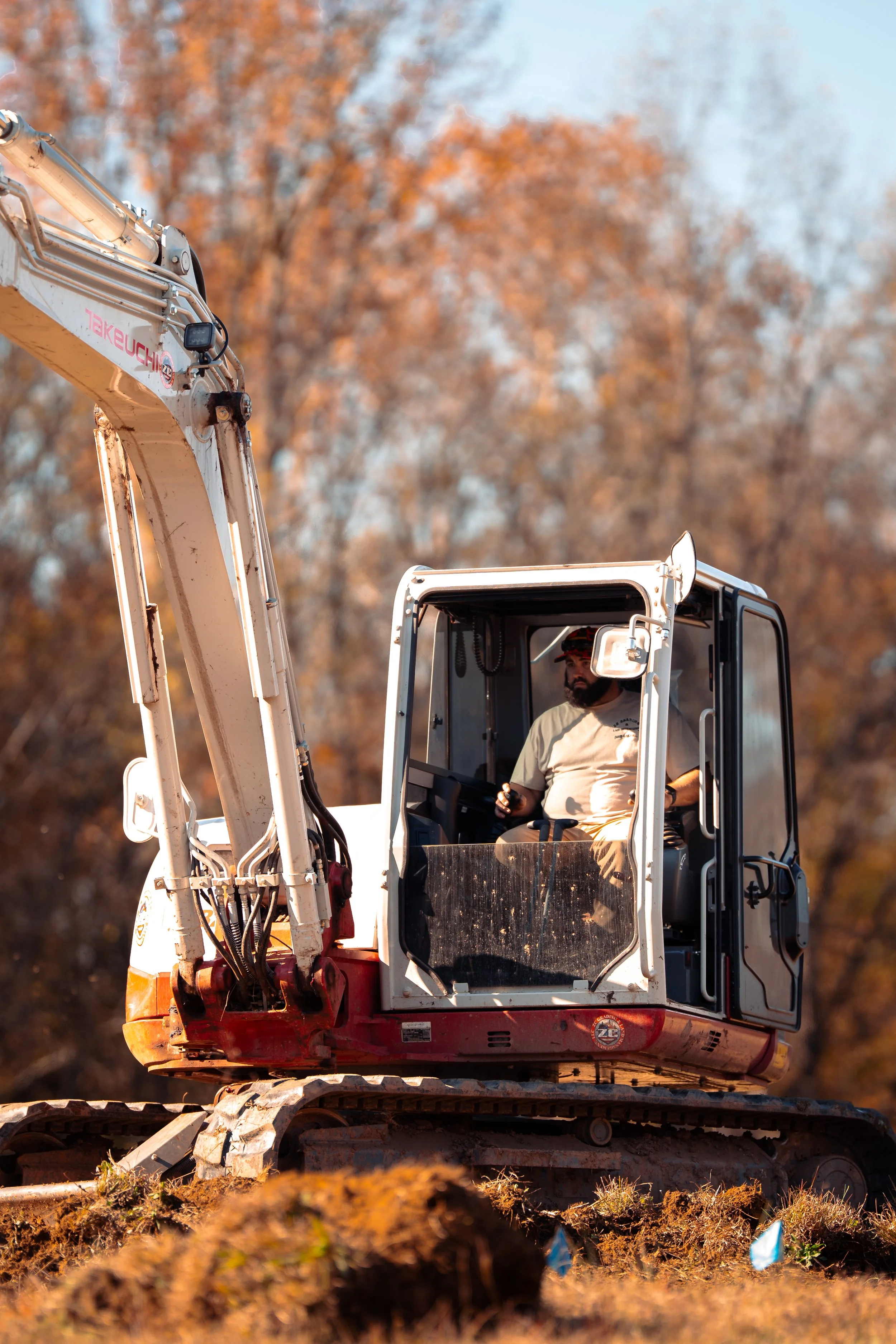 A man operating a small excavator outdoors with autumn-colored trees in the background.
