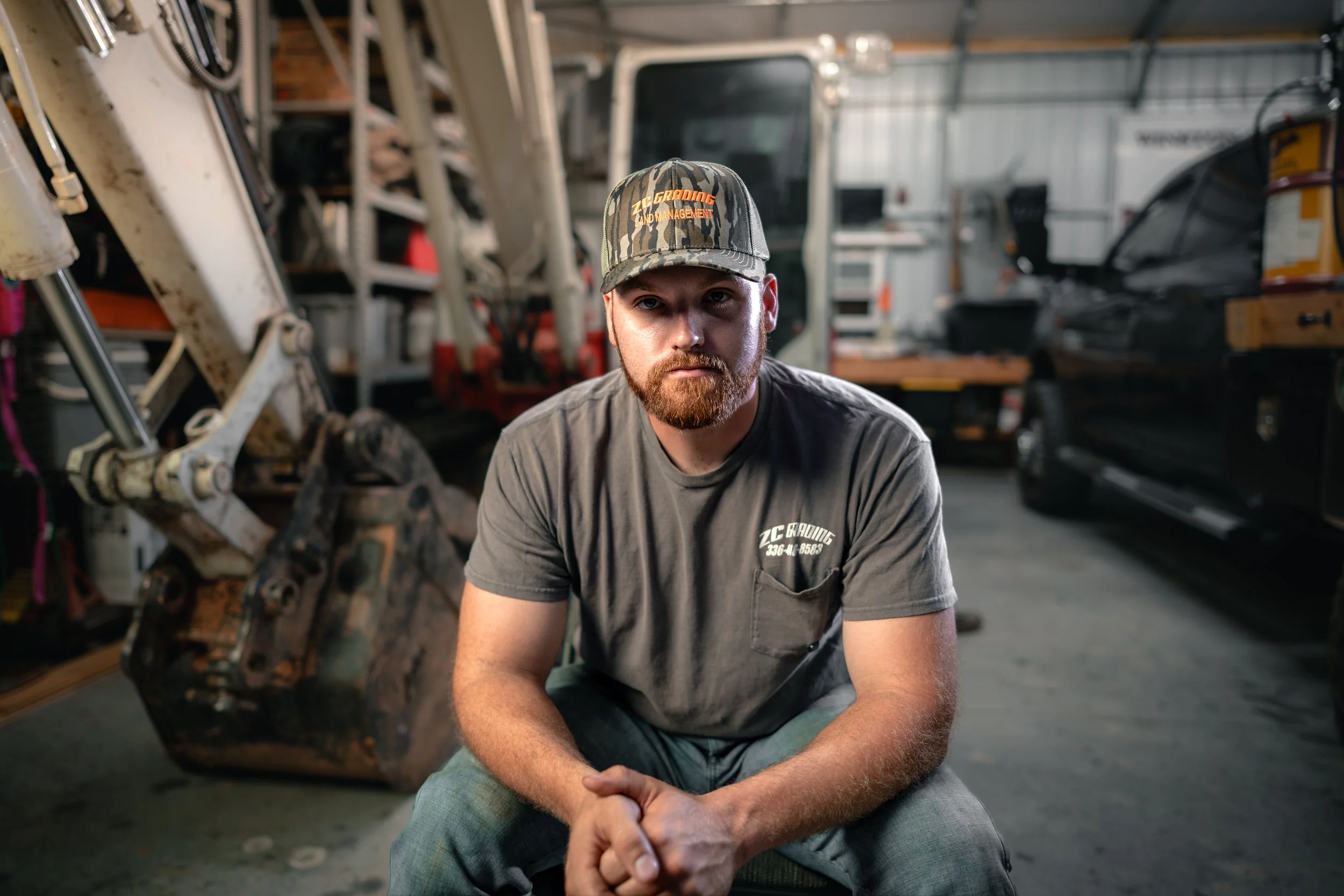 A man with a beard wearing a cap and a t-shirt sitting in a garage or workshop with machinery and a black vehicle in the background.