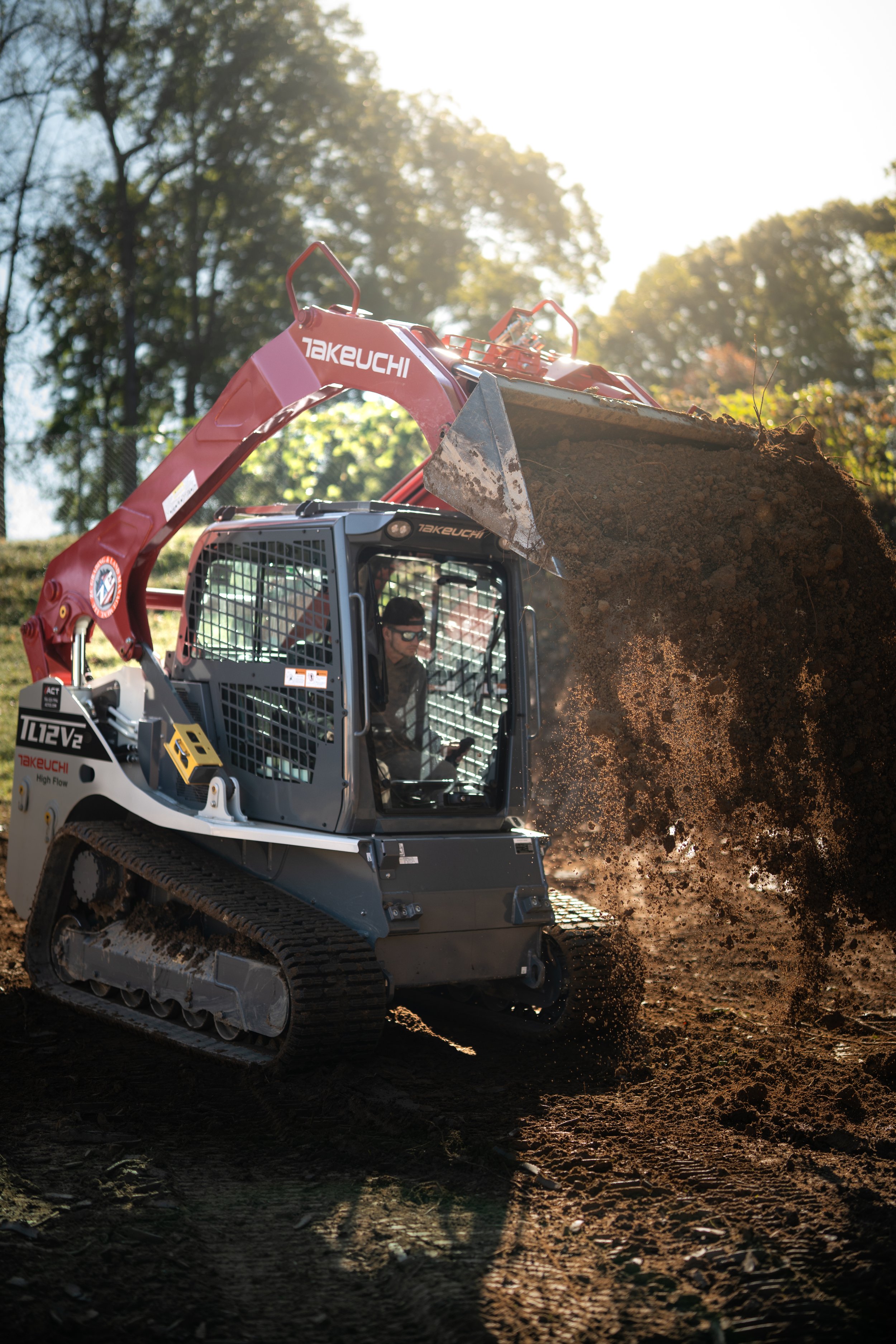 A person operating a compact Takeuchi excavator with a red arm, digging and moving dirt on a construction site with trees in the background.