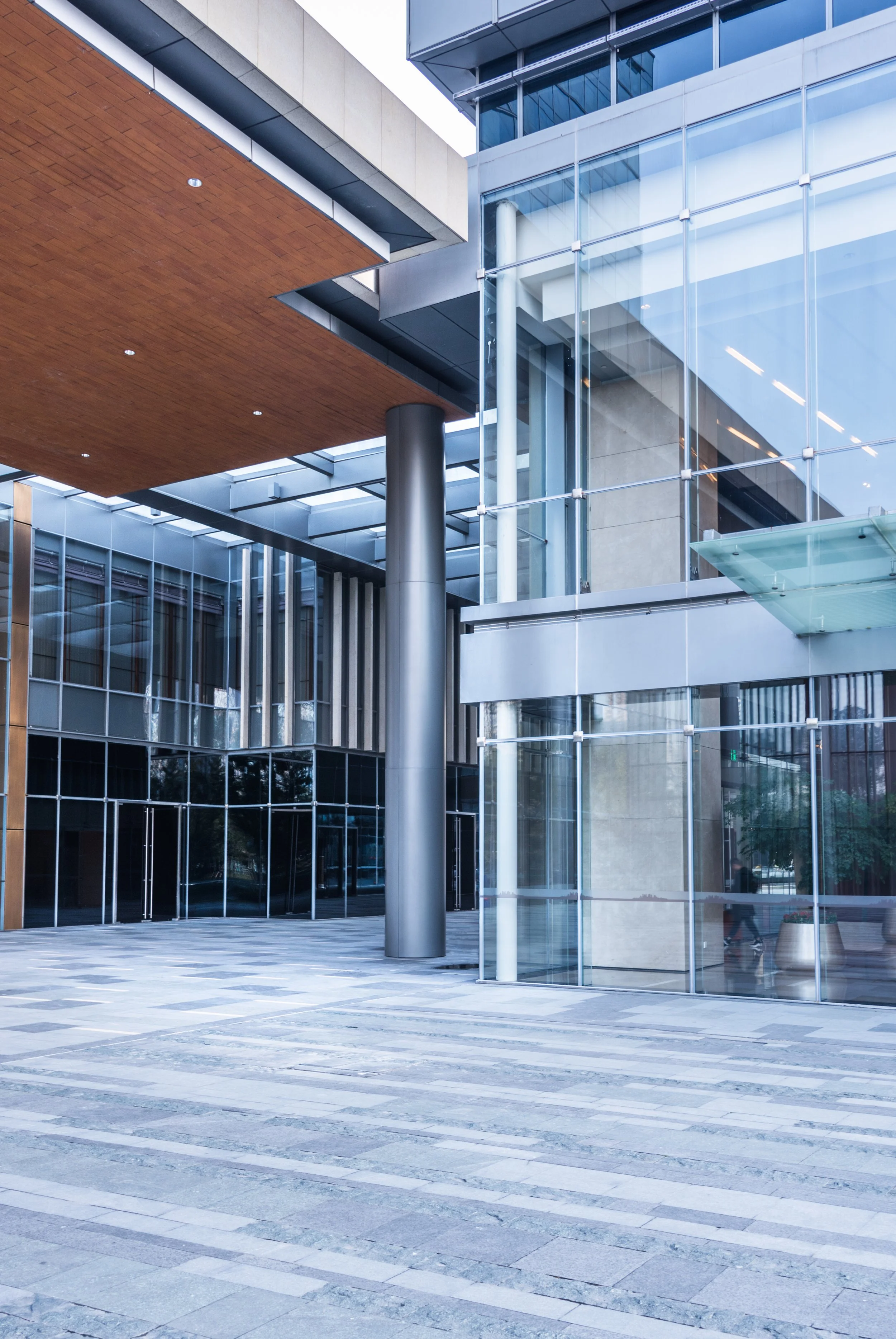 Modern building with exposed structure, glass walls, metal columns, and wooden ceiling overhang, with a paved sidewalk in front.