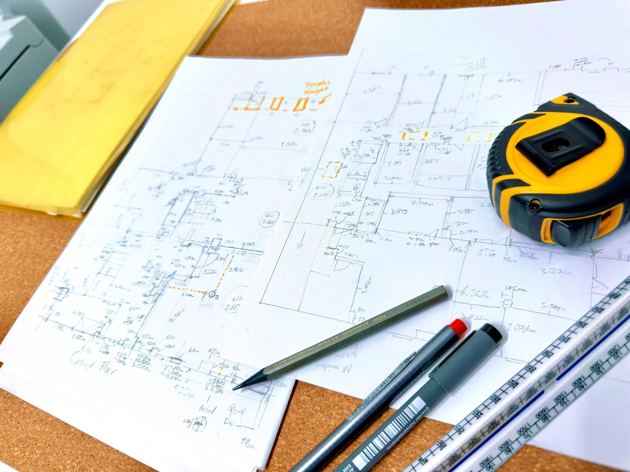 Architectural survey notes on a desk with a yellow folder, a tape measure, markers, a pen, and a scale ruler.