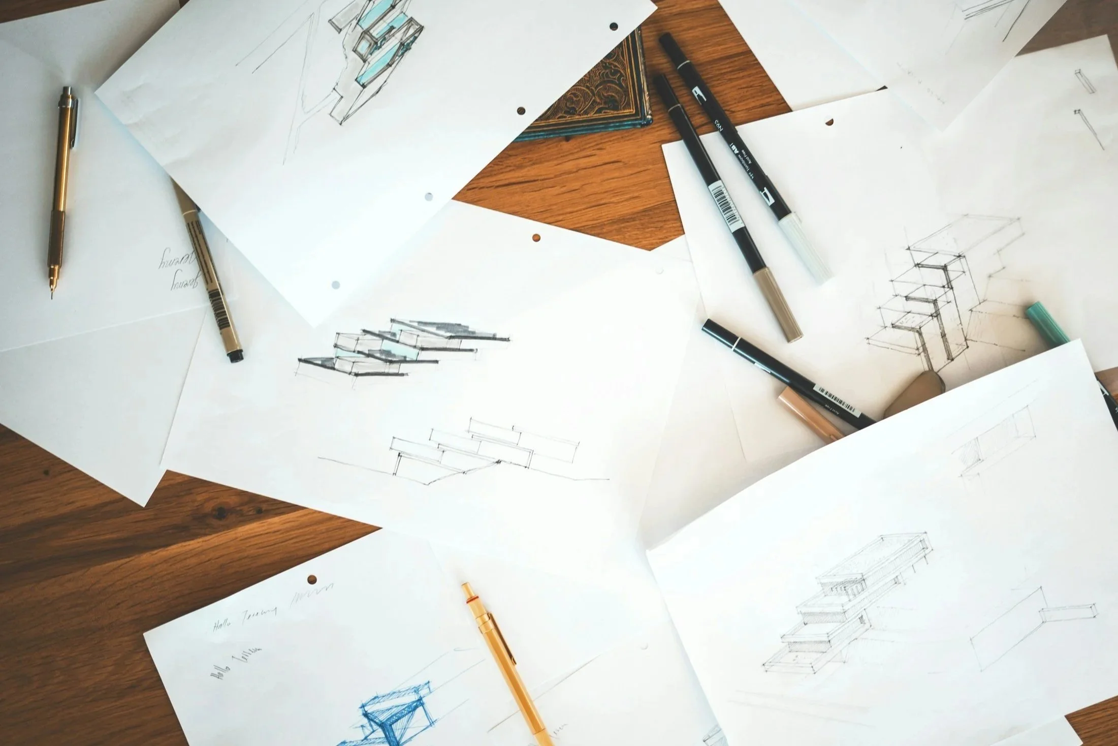 Architectural sketches and plans on white paper scattered on a wooden table, with pens and markers nearby.