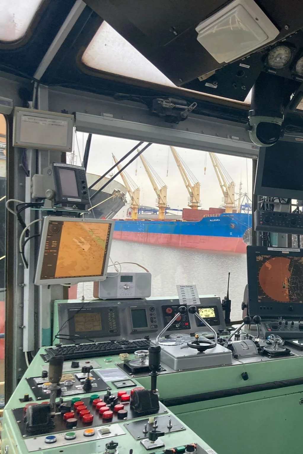 Interior view of a ship's control room showing various navigation and communication equipment, with a large cargo ship and cranes visible through the windows.
