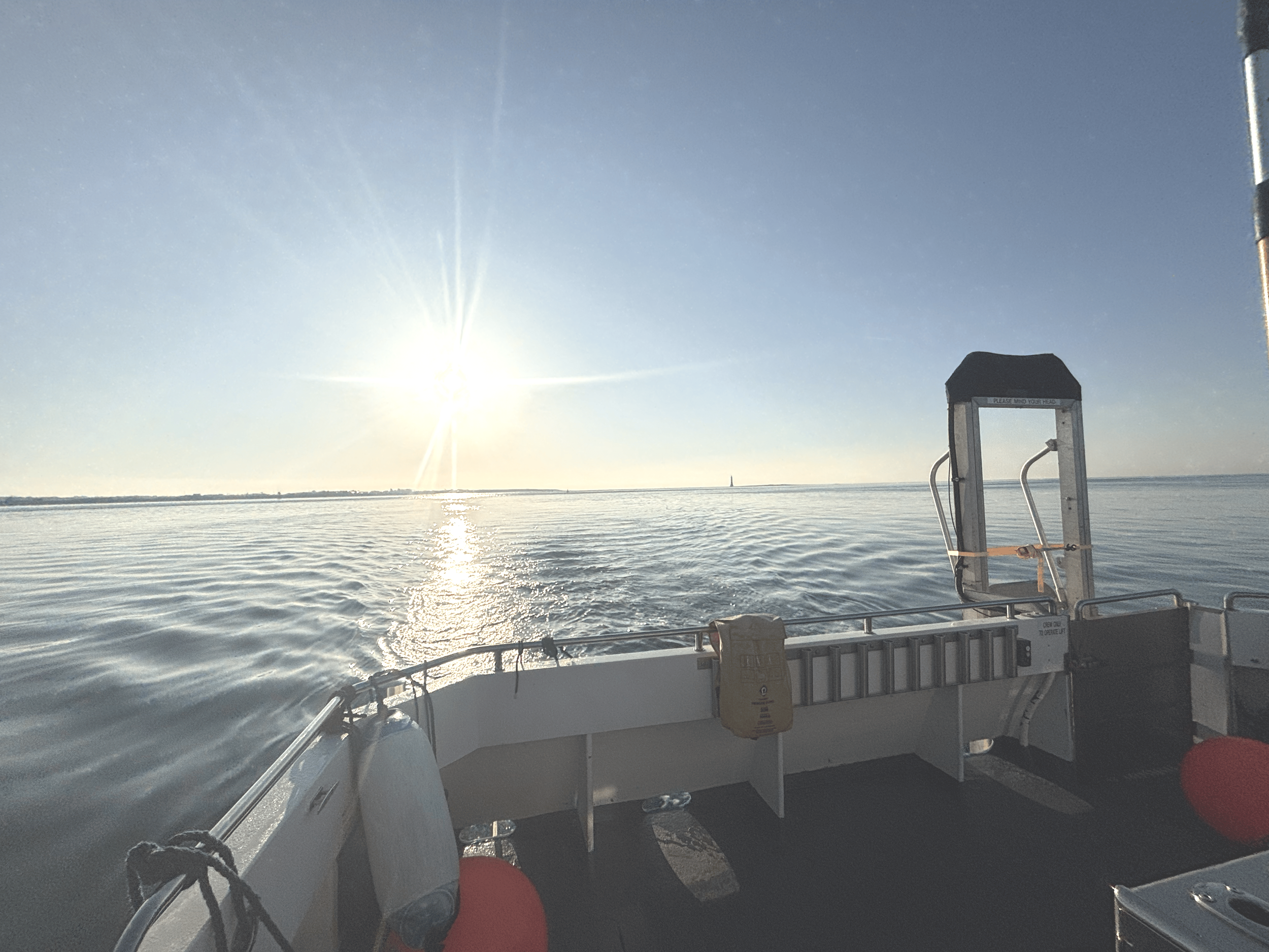 View from a boat on calm water with the sun low in the sky, creating a reflection on the surface. The boat's railing with fenders and a ladder are visible in the foreground.