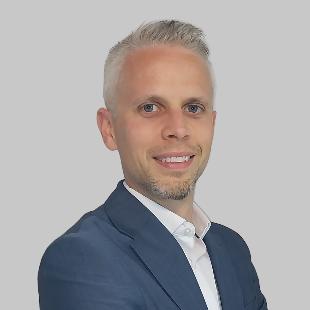 A man with short, silver hair, blue eyes, and a beard, wearing a dark blue suit jacket and a white collared shirt, smiling against a plain light gray background.