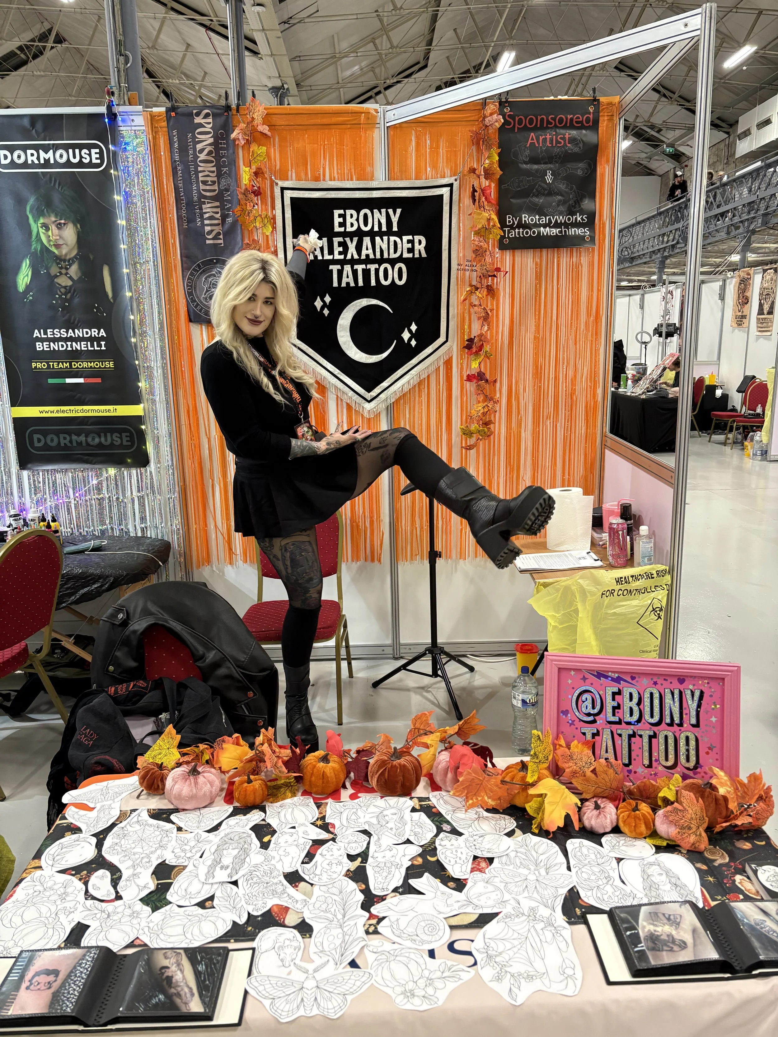 A woman with blonde hair, black clothing, and tattoos is standing at a booth for Ebony Alexander Tattoo. She is lifting her leg in front of a colorful display with signs advertising her tattoo art. The booth has fall-themed decorations including small pumpkins and autumn leaves on a table with tattoo sketches and artwork, at a convention or event.