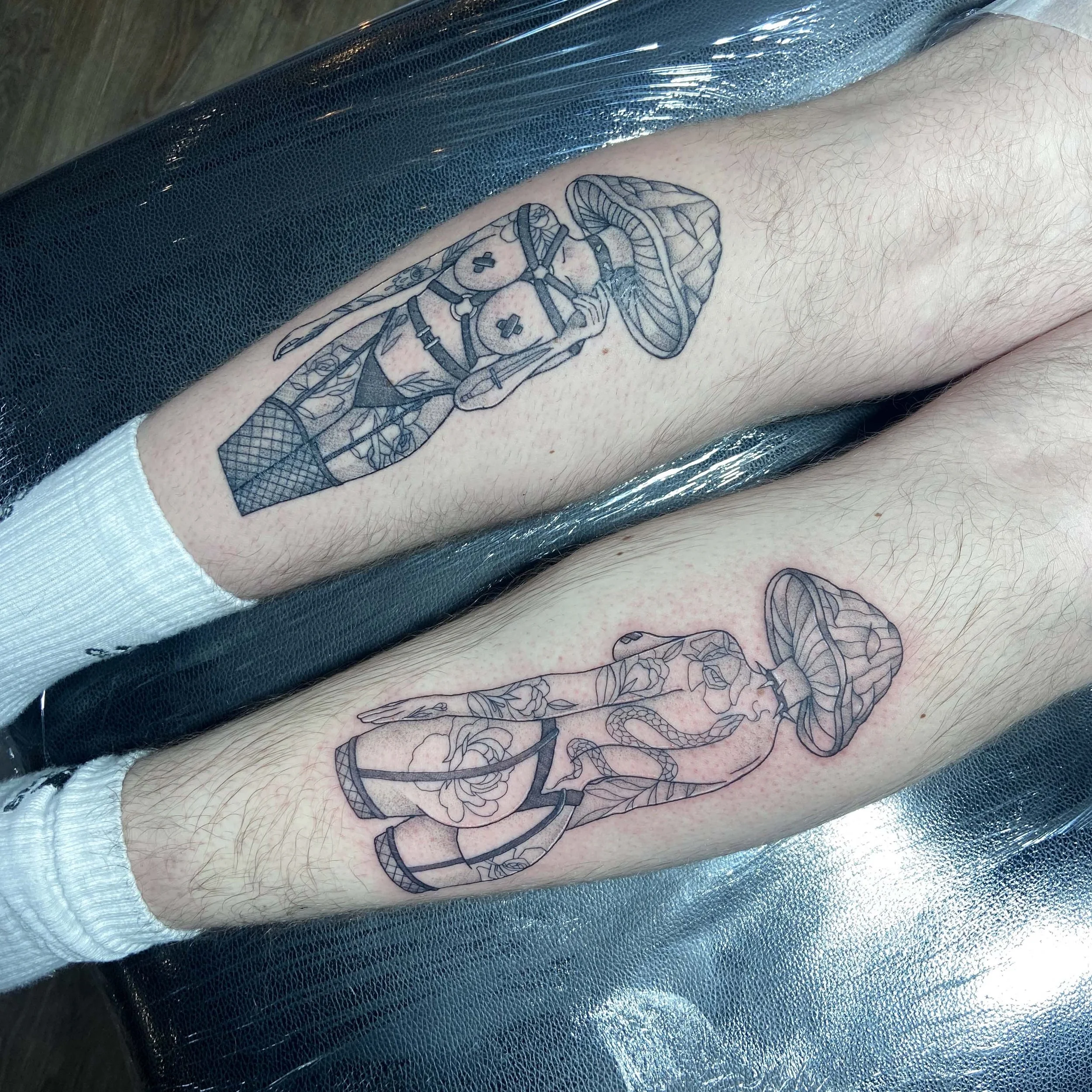 Two person's legs with matching black outline mushroom tattoo designs featuring snakes and roses.