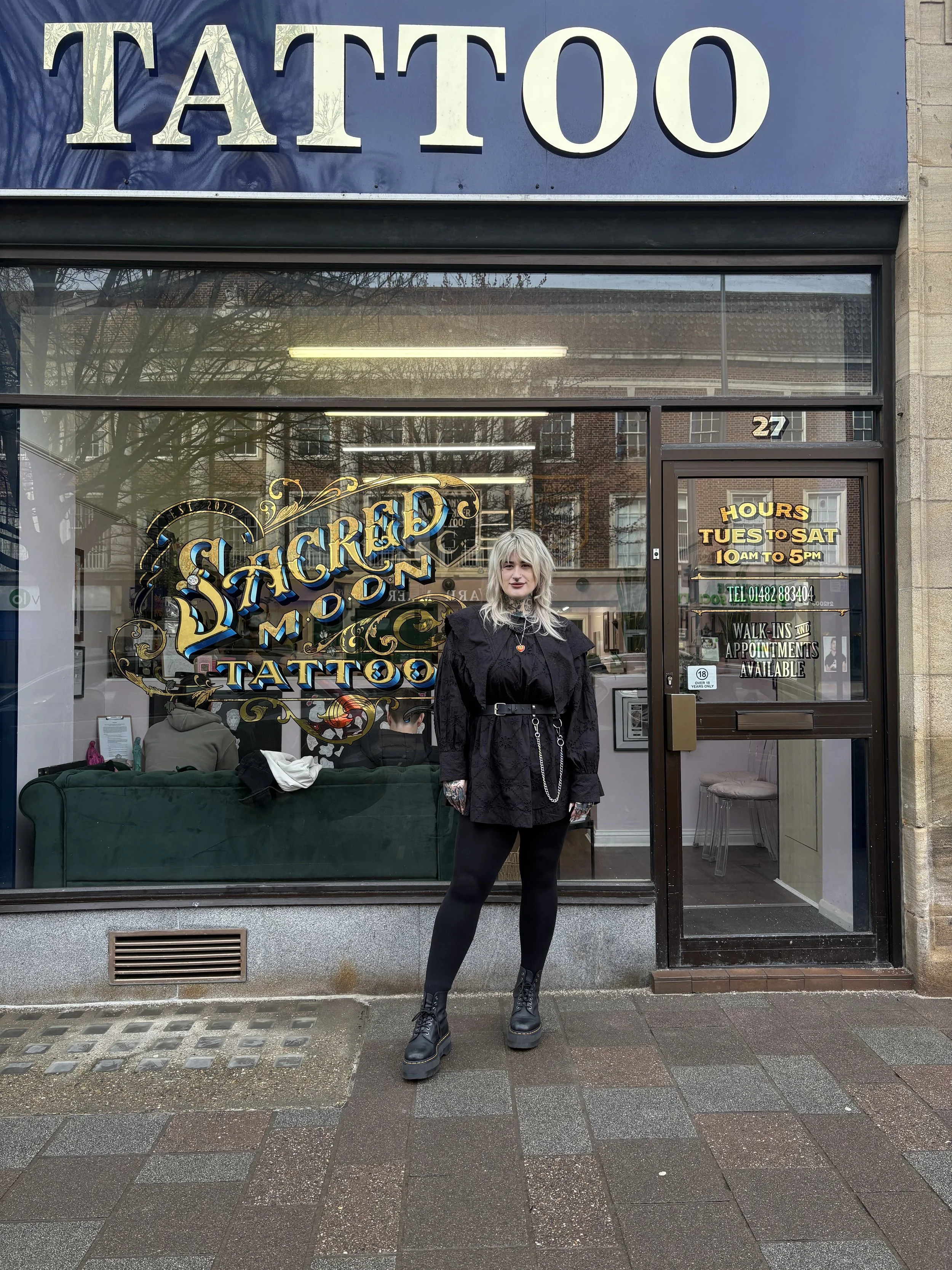 Woman standing outside Sacred Moon Tattoo shop, wearing a black jacket, black leggings, and combat boots.