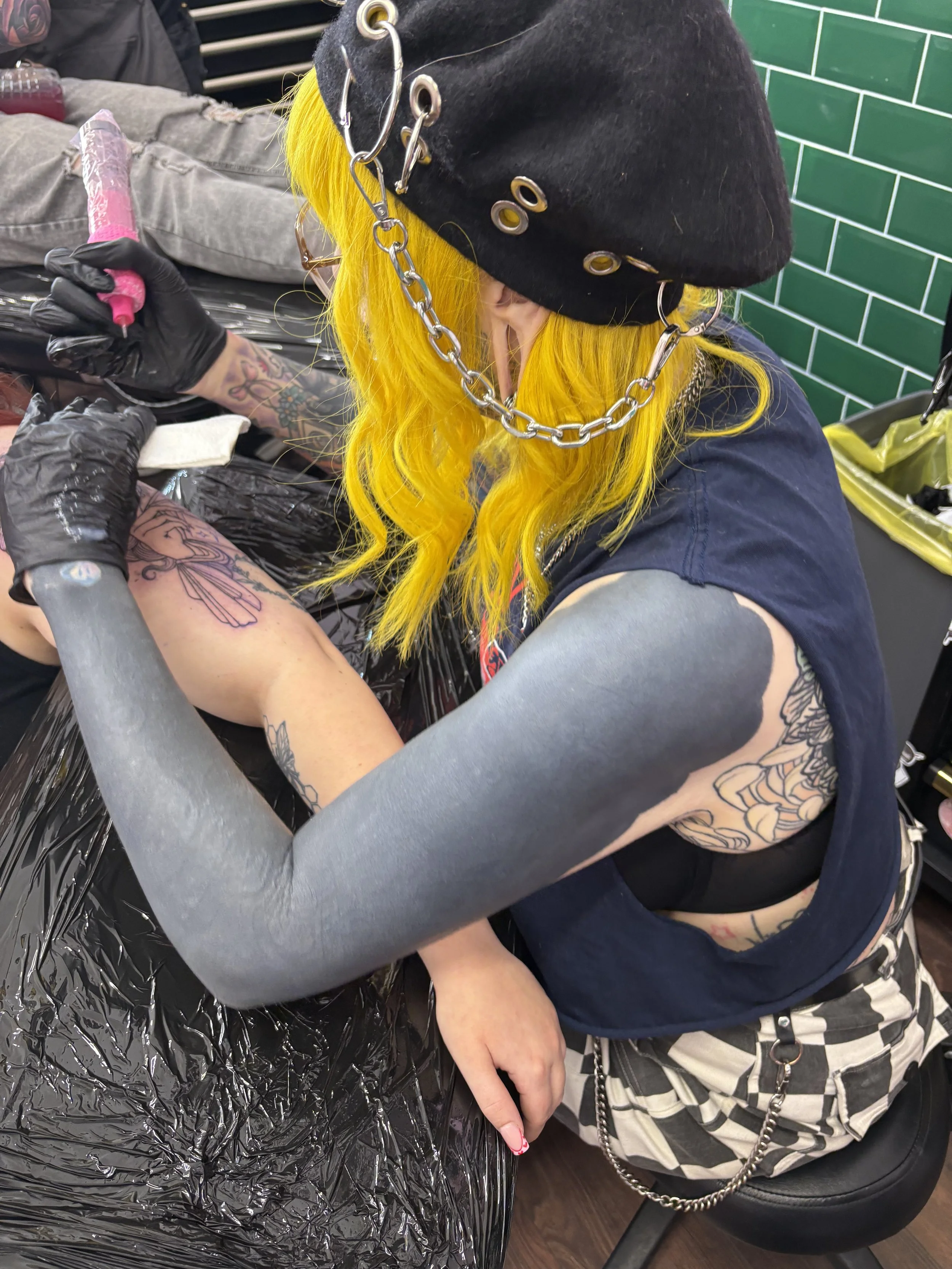 A tattoo artist with bright yellow hair, wearing a black beanie with metal eyelets and a chain, is tattooing a person's arm. The artist has tattoos on her arms and is wearing black gloves. The person receiving the tattoo has tattoos on their arm and is sitting with their hand resting on a plastic-covered surface in a tattoo studio with green tile walls.