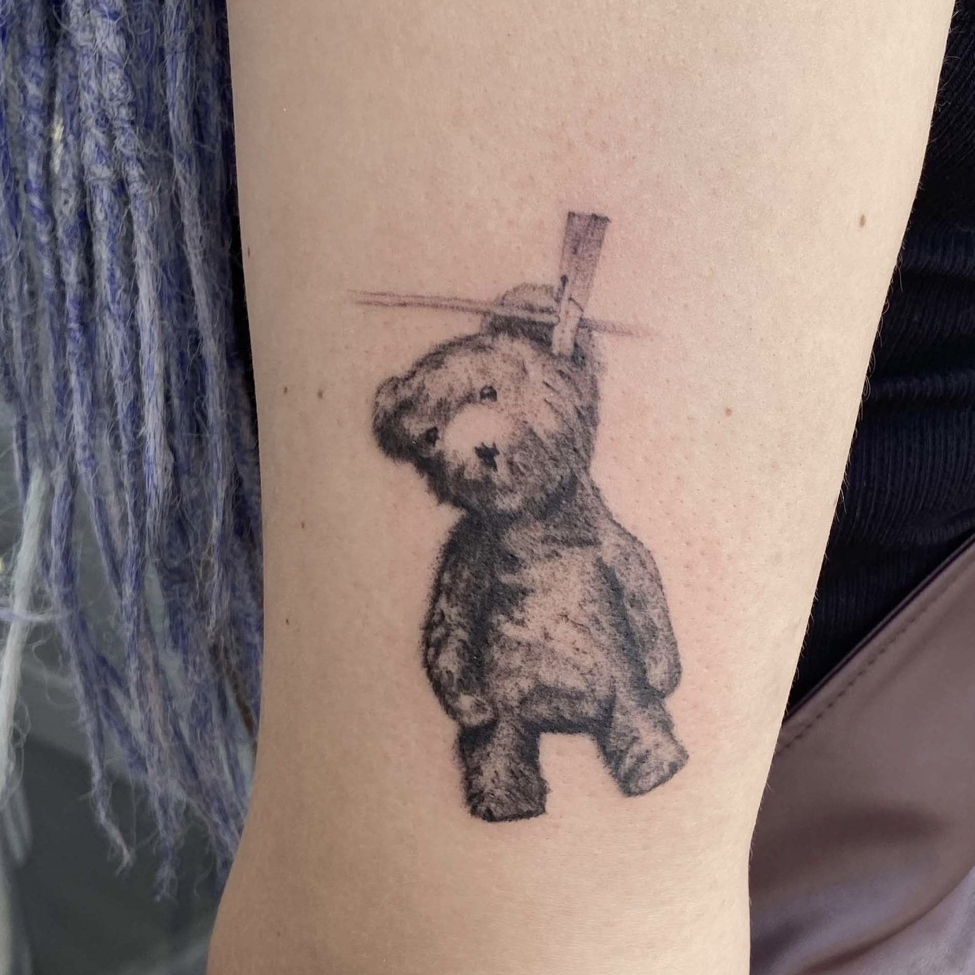 Tattoo of a teddy bear hanging from a clothesline by one paw with a clothespin.