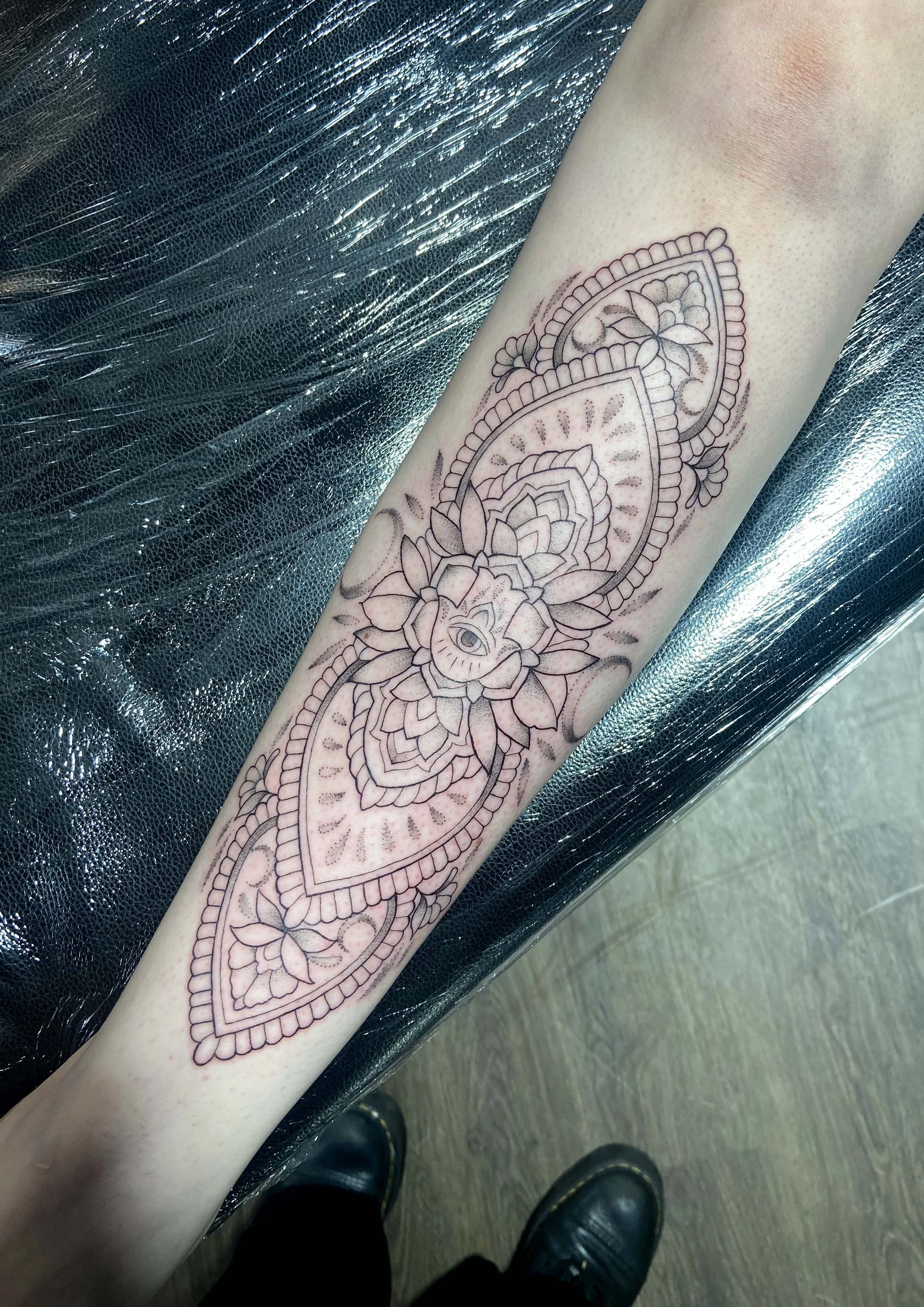 Line art tattoo of a mandala with floral and eye details on someone's forearm.