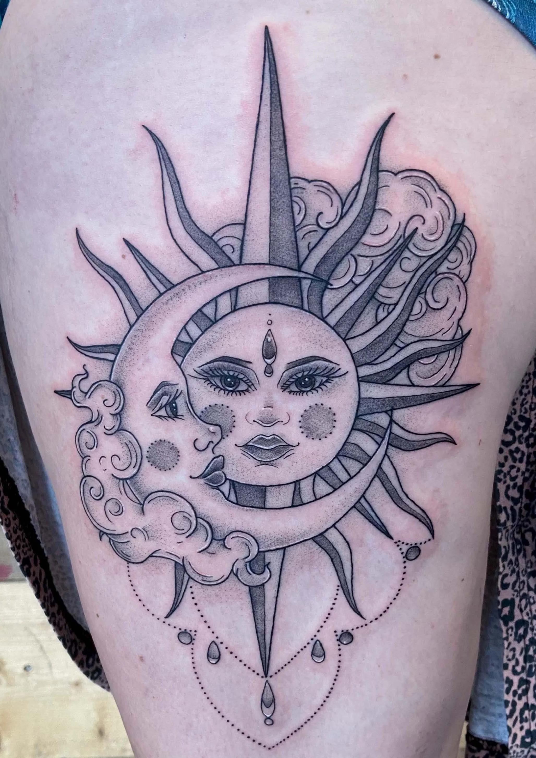 A tattoo of a celestial scene featuring a sun, moon, and a face with detailed eyes and lips. The sun has rays extending outward, the moon is crescent-shaped, and both are surrounded by clouds and decorative dots with hanging jewelry-like accents.