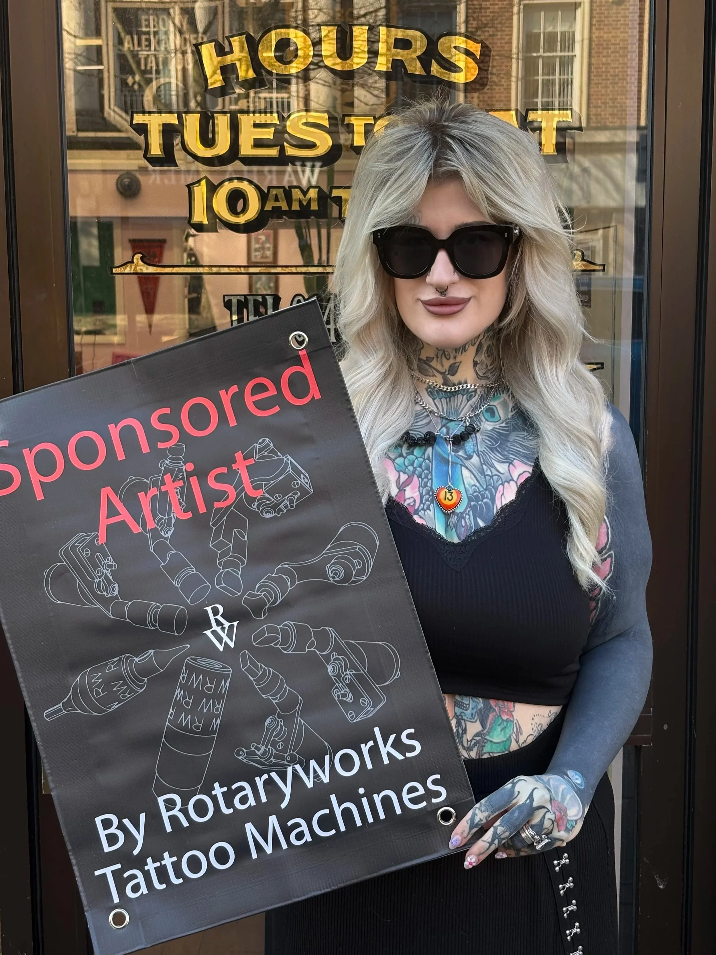 A woman with long, wavy blonde hair, wearing large black sunglasses, a septum nose ring, and multiple tattoos, holding a sign that reads 'Sponsored Artist By Rotaryworks Tattoo Machines'. She is standing outside a storefront with a sign indicating hours of operation, with the hours visible in the background.