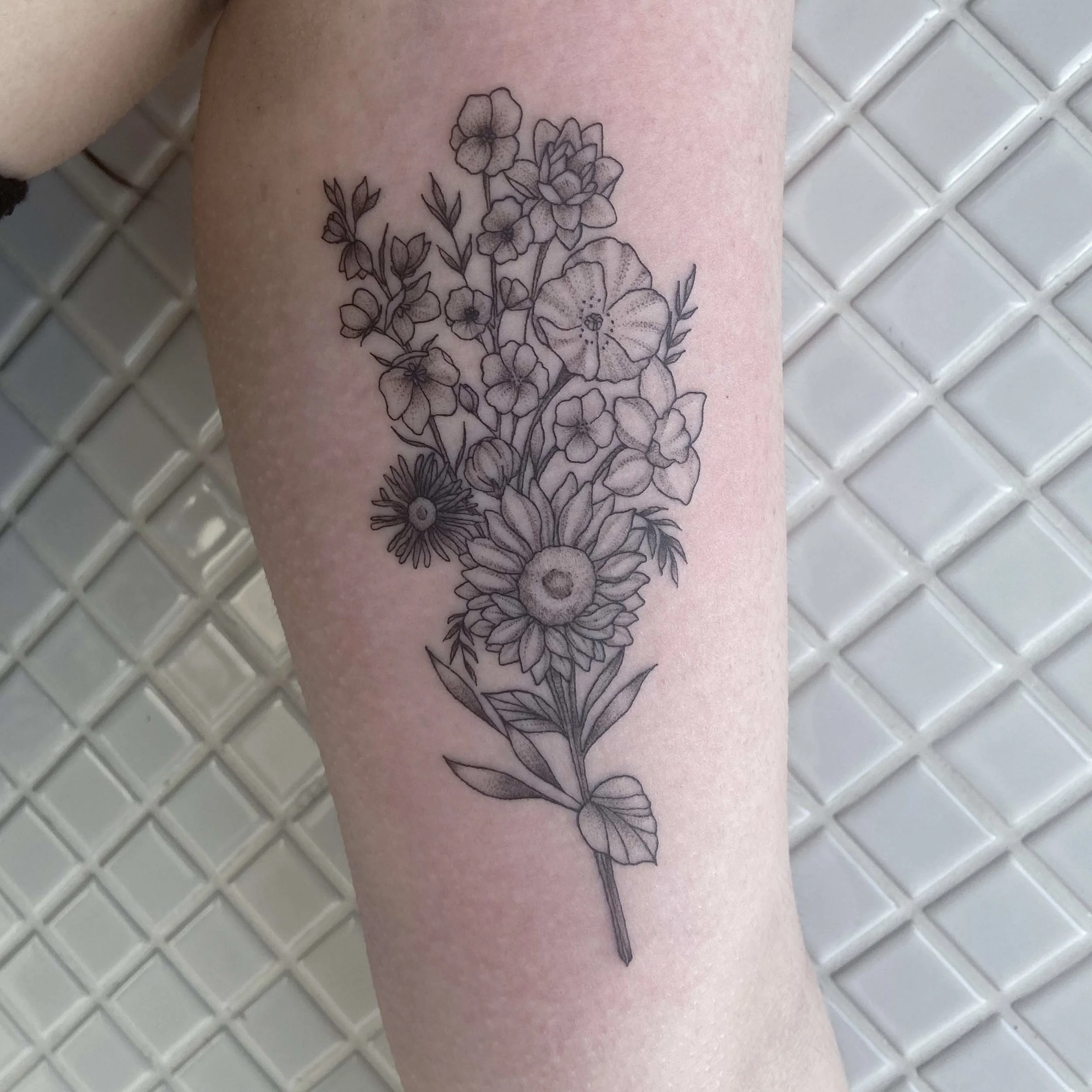 Black and gray floral tattoo on a person's forearm, featuring a bouquet of various flowers including daisies, poppies, and other blossoms with detailed leaves and stems.