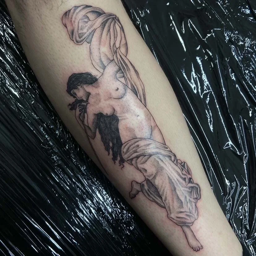 Tattoo of a reclining nude woman with long dark hair, draped in cloth, on a person's forearm.