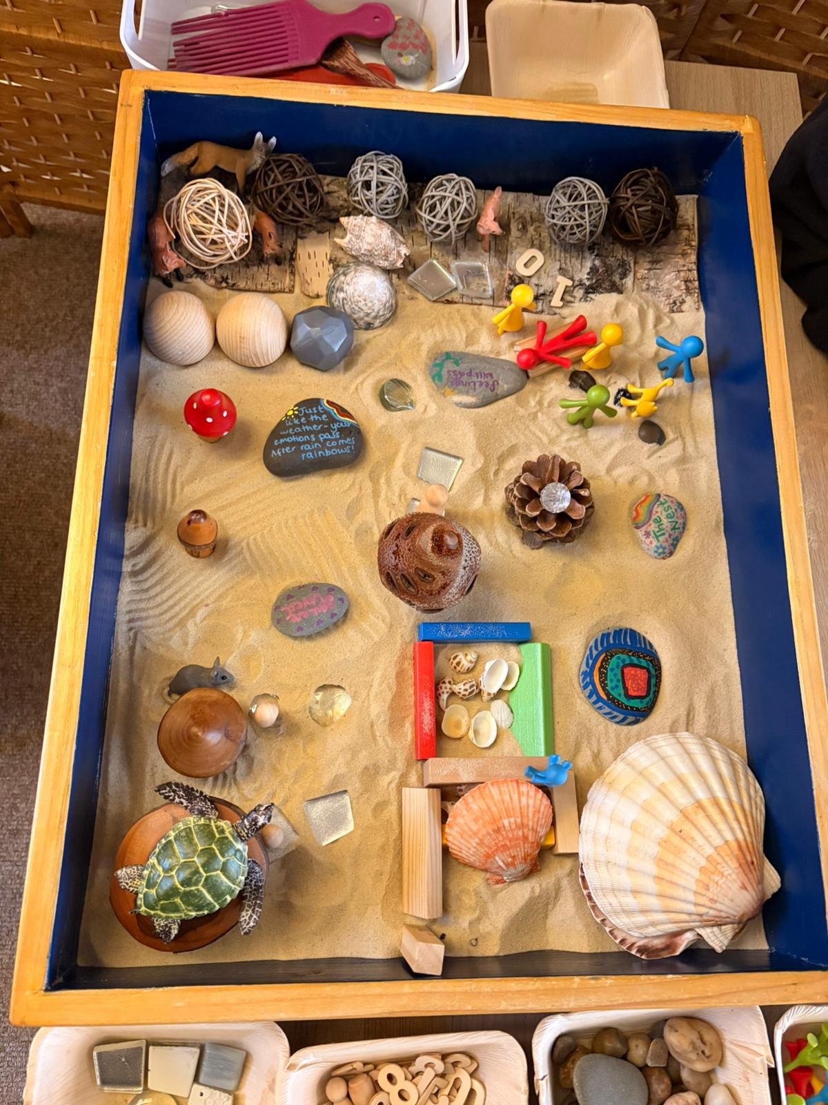 A sensory or play box filled with sand, seashells, miniature figurines, painted rocks, small toys, a turtle figurine, and decorative items, likely used for tactile exploration and play.
