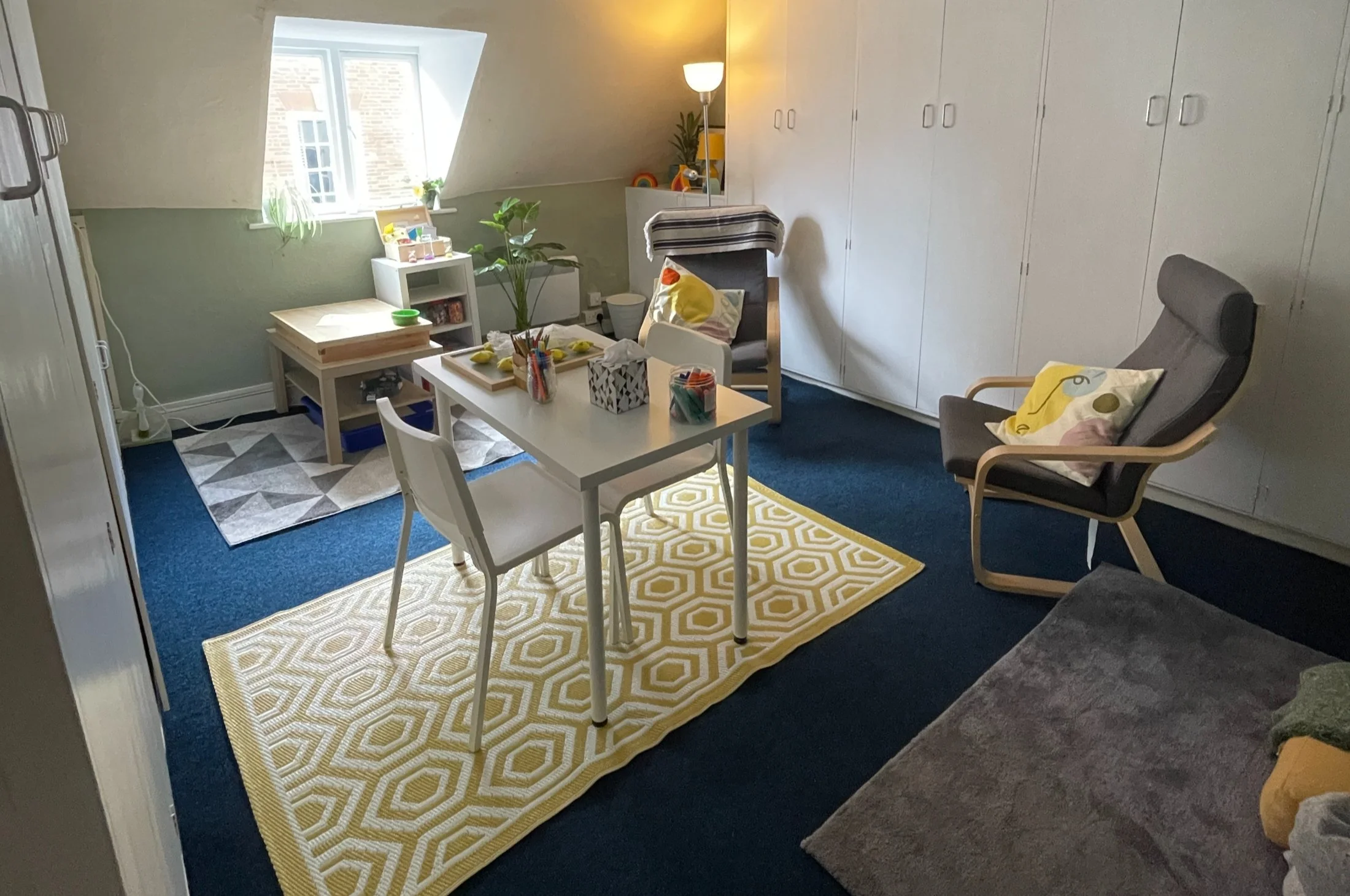 A cozy room with a white table and two chairs, a yellow patterned rug, a modern gray armchair with a pillow, and a blue carpet. There are shelves and cabinets, a window with sunlight, houseplants, and various items on tables and shelves.