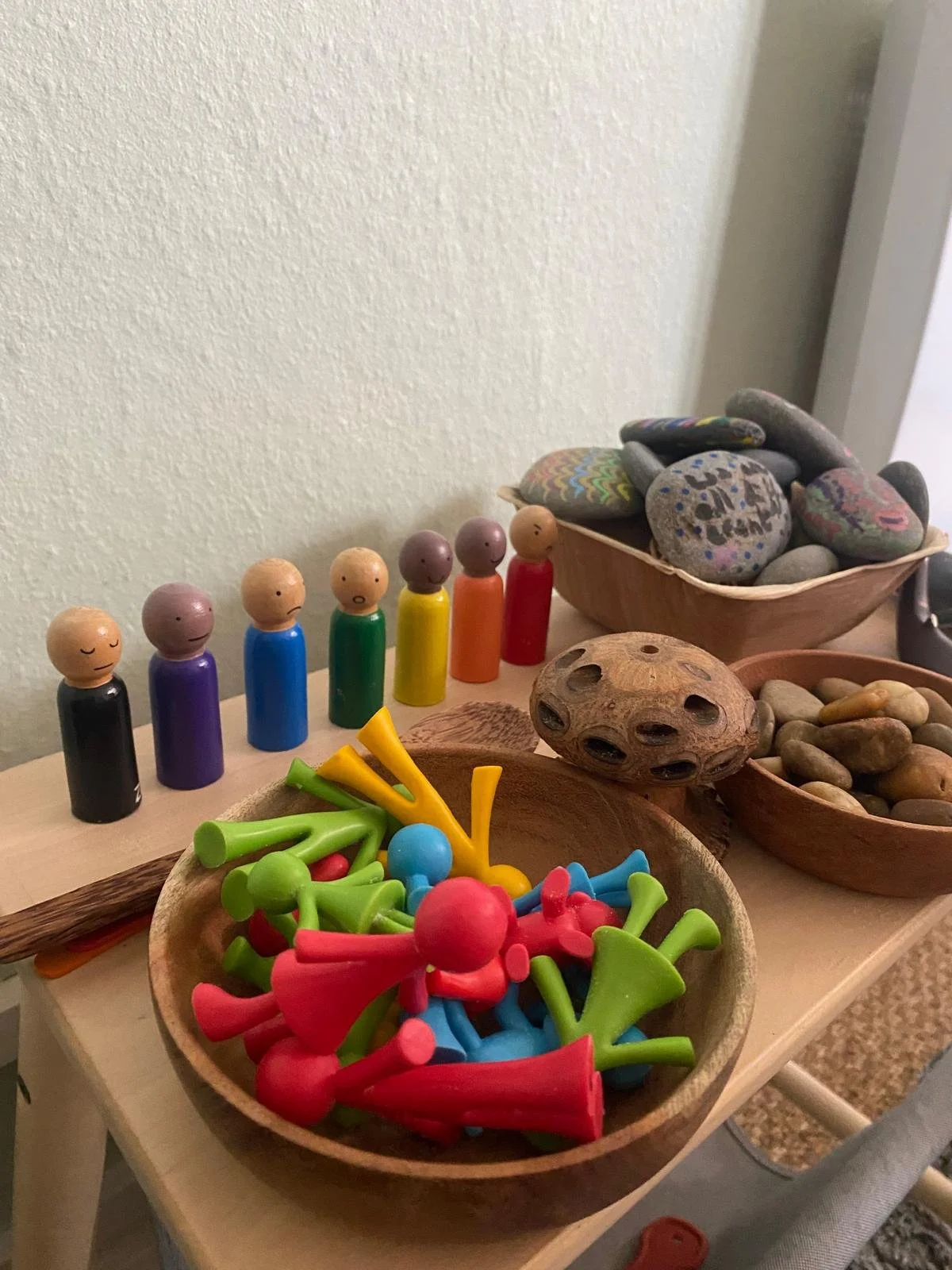 Colorful wooden peg dolls, painted rocks with various designs, and small plastic figurines in bowls and on the table.