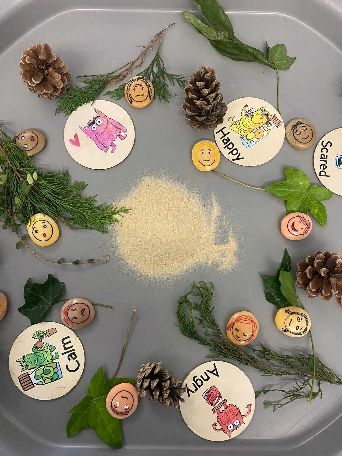 A collection of painted rocks with smiling faces, pinecones, green leaves, and sprigs, arranged in a circle on a gray surface.