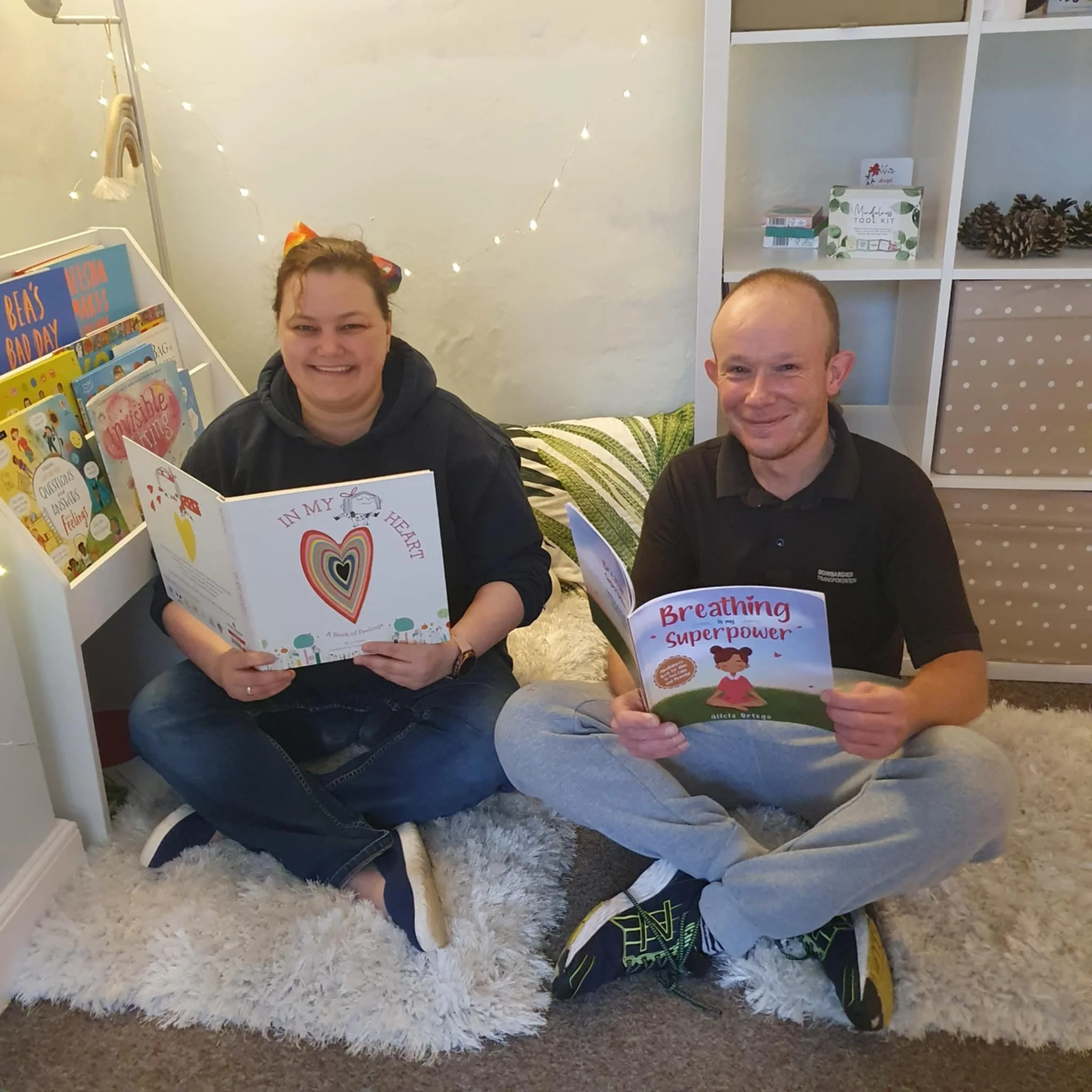 Two people sitting on a fluffy white rug reading children's books. The person on the left is holding a book titled 'In My Heart,' and the person on the right is holding a book titled 'Breathing is my Superpower.' They are in a cozy room with bookshel