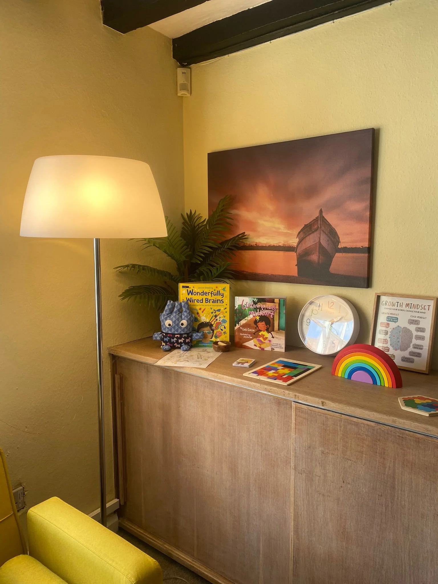 A sideboard with children's books, a stuffed owl toy, decorative rainbow, clock, framed diagrams, potted plant, and a large sunset boat photograph. Yellow armchair in foreground, standing lamp to left.