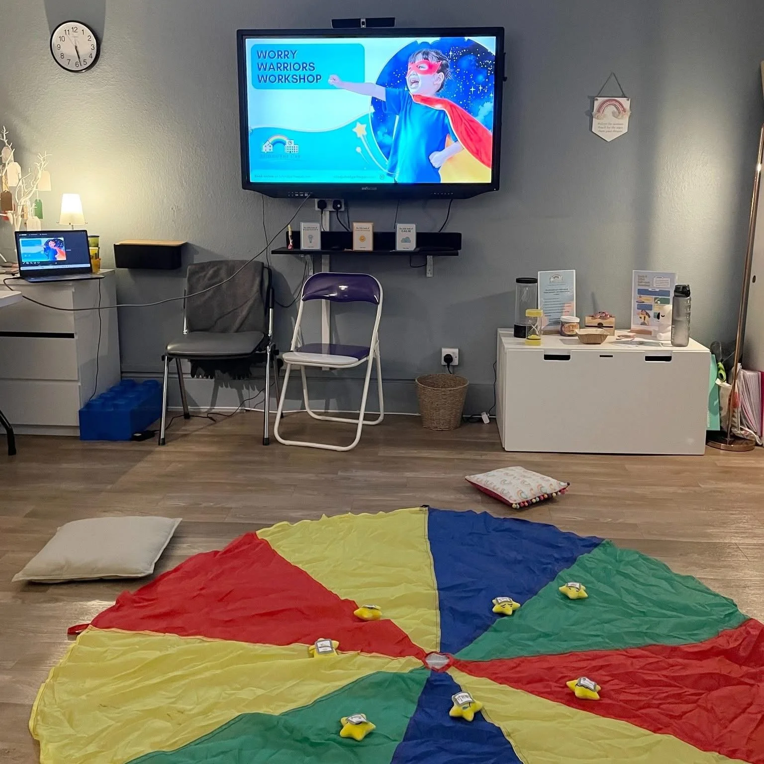 A room set up for a children's workshop or activity with a colorful parachute on the floor, pillows, a large television screen displaying a cartoon superhero, a laptop, chairs, and various informational materials.