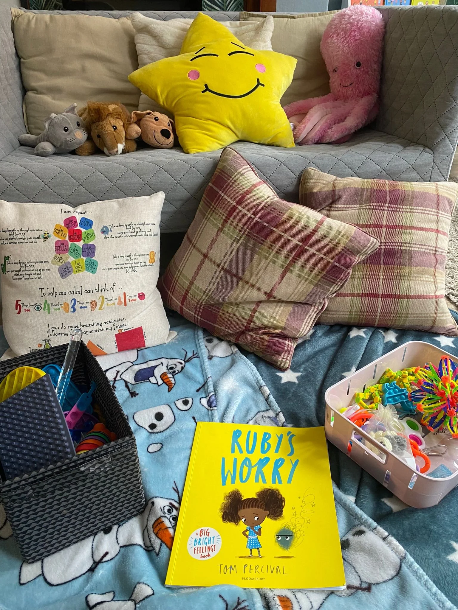Child's cozy play area with stuffed animals, pillows, a book titled 'Ruby's Worry', a box of colorful craft supplies, and a blanket featuring Olaf from Frozen.