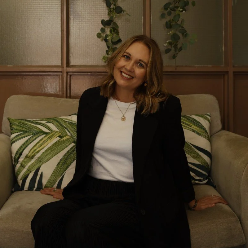 A woman with shoulder-length blonde hair, wearing a black blazer over a white top, sitting on a beige couch with green and white patterned pillows, smiling at the camera in a cozy indoor setting with wooden and frosted glass wall panels in the background.