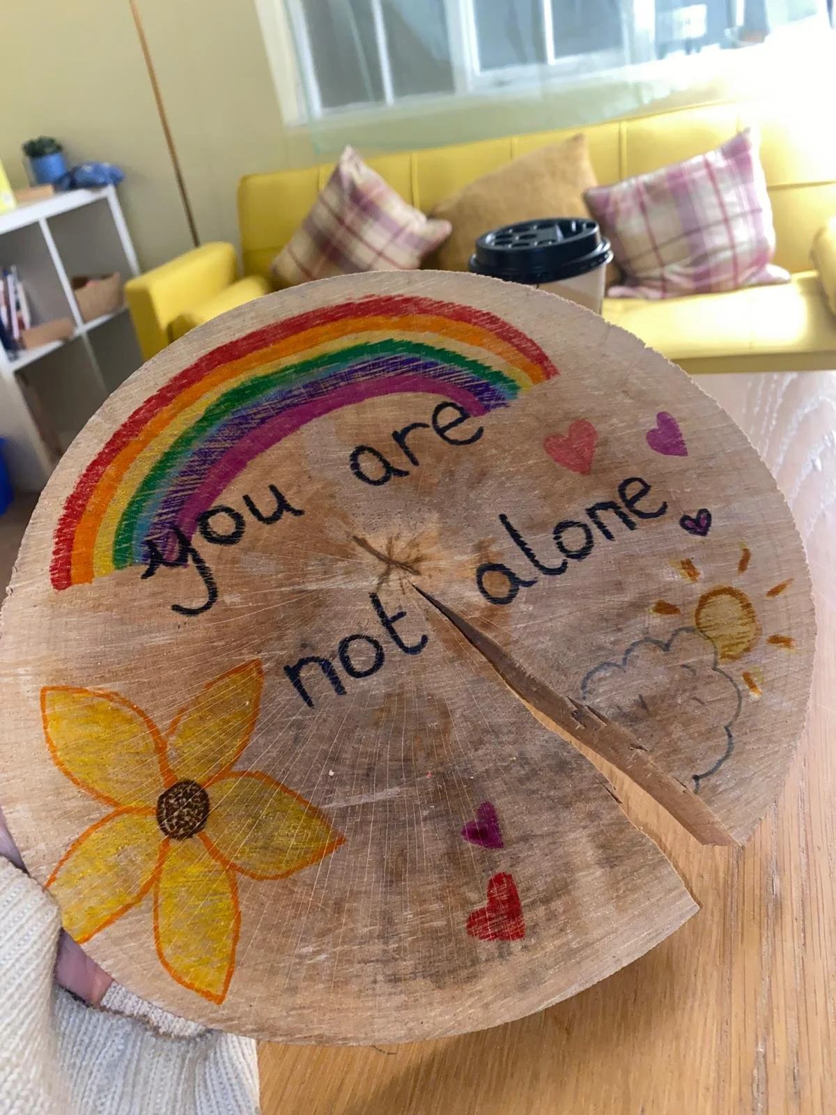 Decorative wooden slice with a rainbow, flower, hearts, sun, cloud, and the phrase 'you are not alone' written on it.