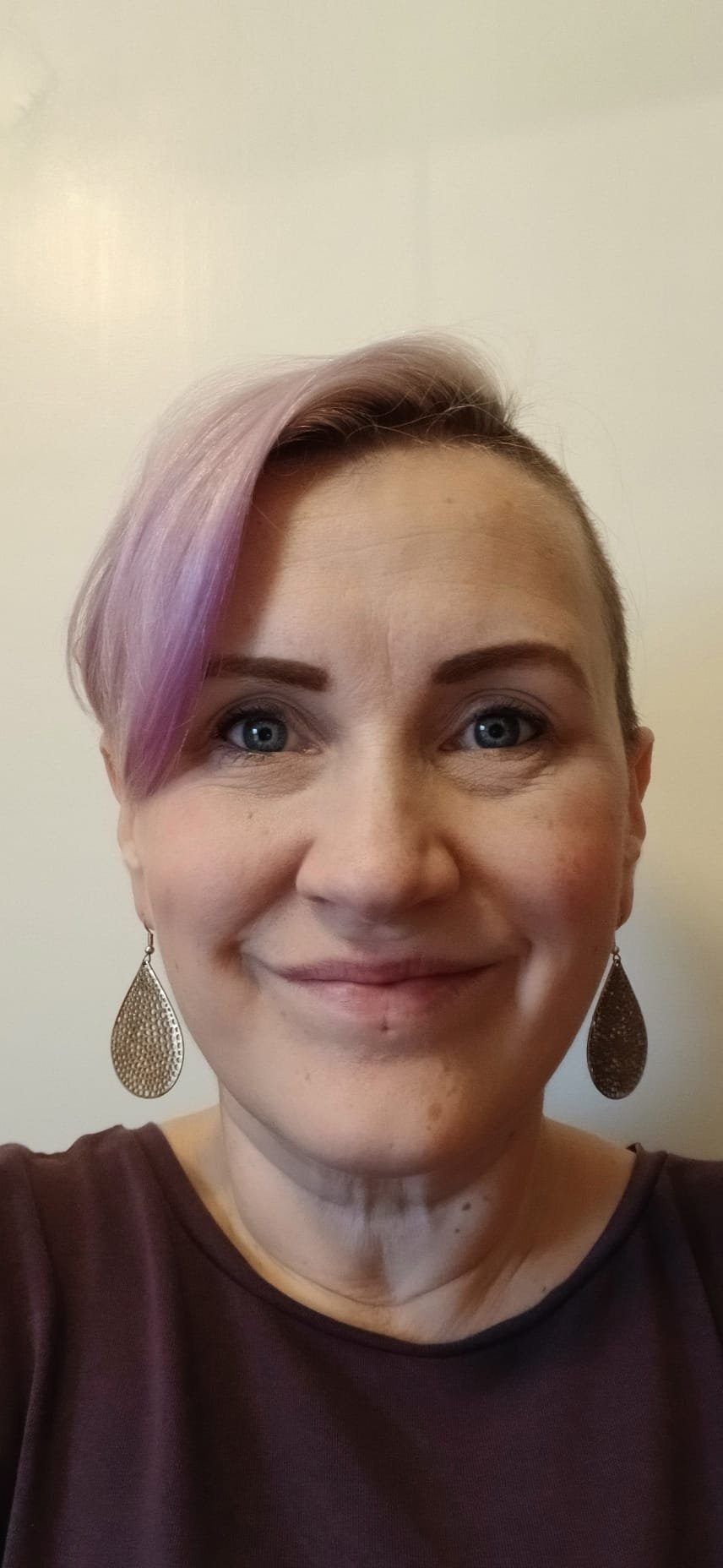 Close-up selfie of a woman with short hair, with a purple streak, wearing large teardrop earrings and a dark top, smiling against a plain light background.