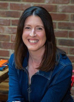 Portrait of a woman with shoulder-length dark hair, smiling, wearing a blue jacket, standing in front of a brick wall.