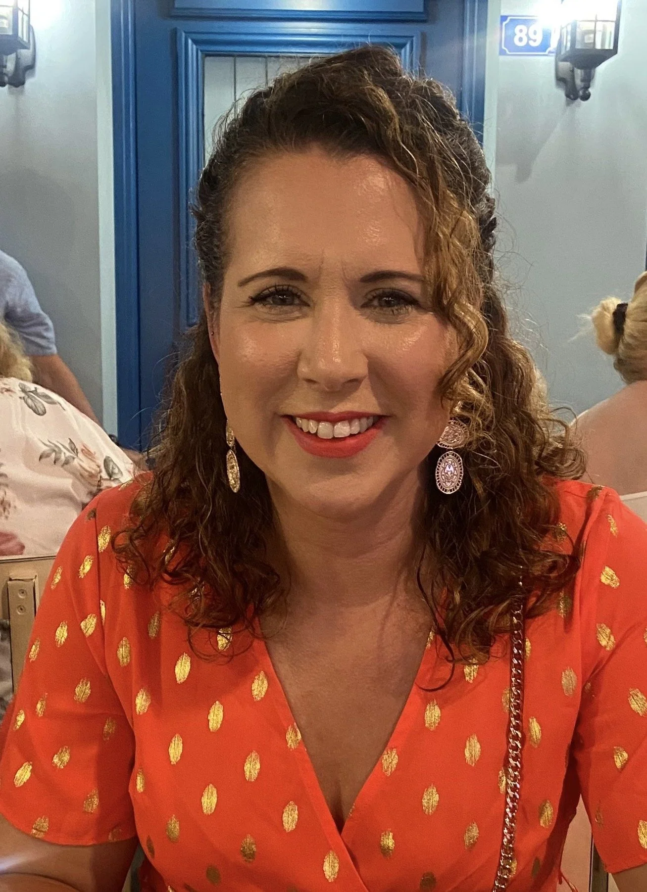 A smiling woman with curly brown hair, wearing an orange dress with gold polka dots, and large dangling earrings, sitting in a restaurant or event space with other people visible in the background.