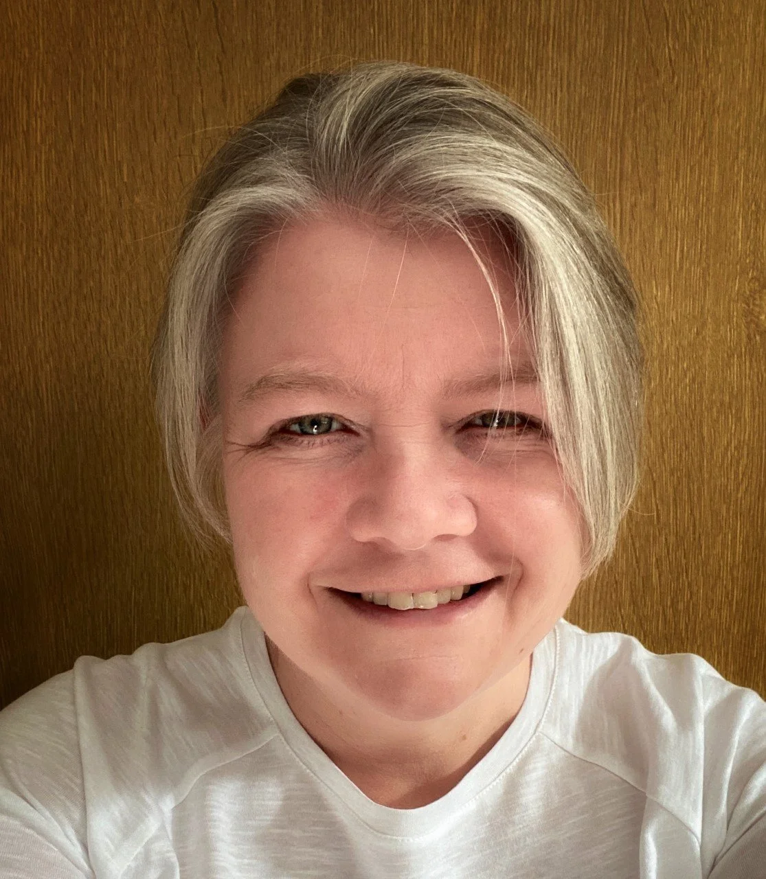 A smiling woman with short blonde hair and light blue eyes, wearing a white shirt, standing in front of a wooden background.