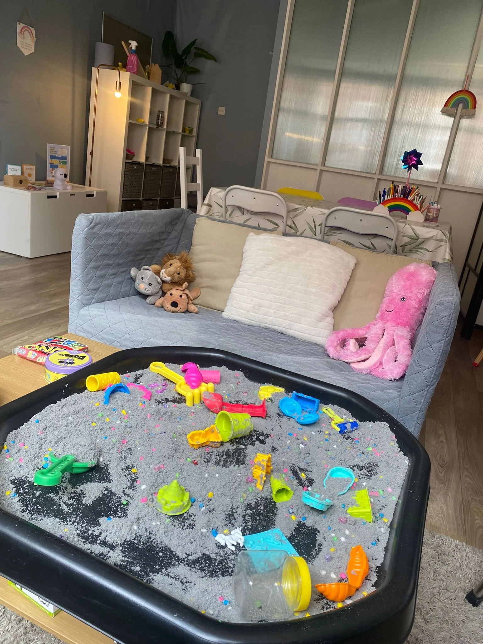 A sensory play table filled with sand and colorful toys, situated in a cozy living room with plush pillows, a sofa, and children's decorations.