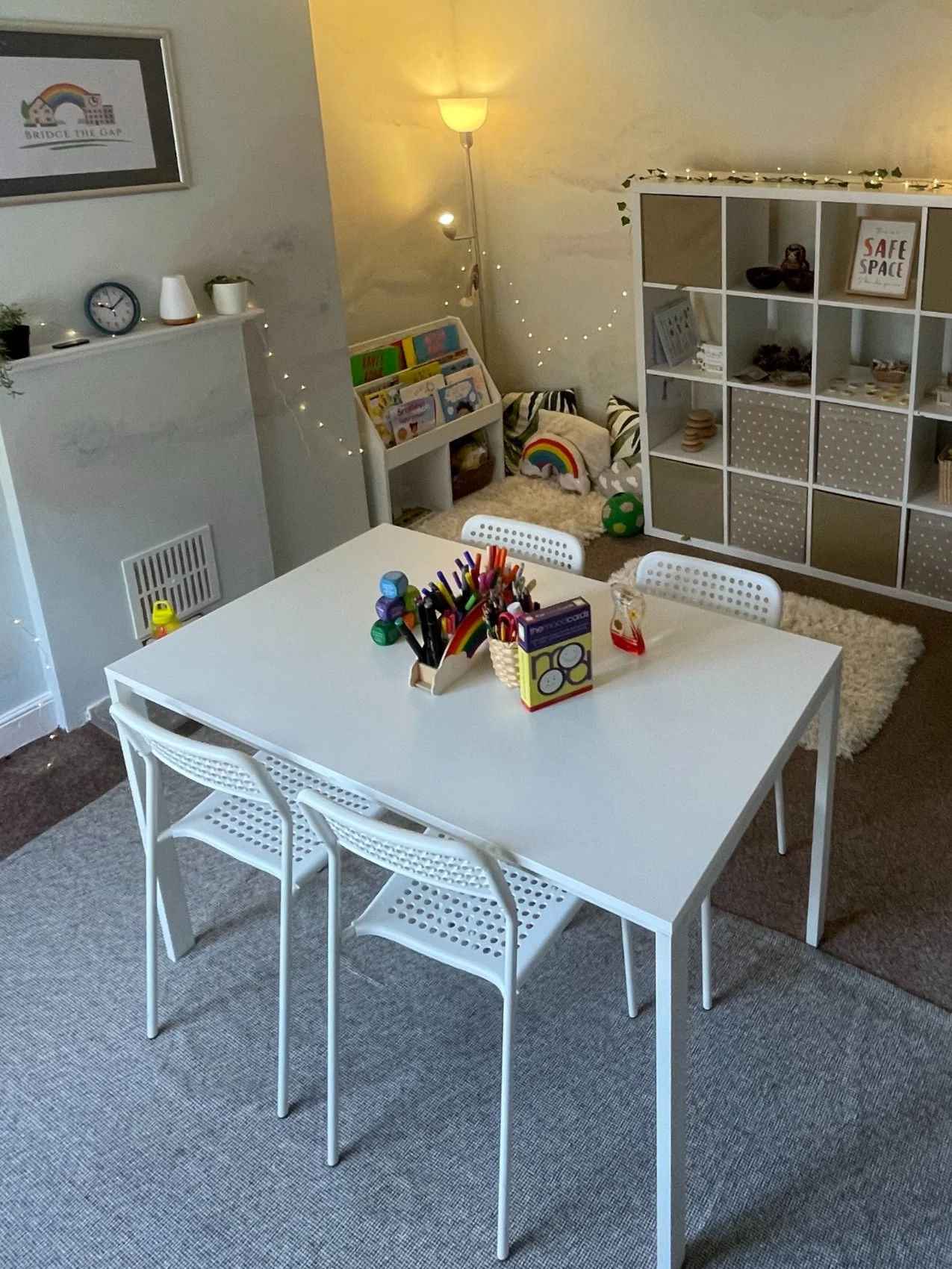 A children's playroom with a white table and three matching chairs, with markers and supplies on top; a corner with books, pillows, and toys; a wooden bookshelf with storage boxes; paper posters and fairy lights on the walls.