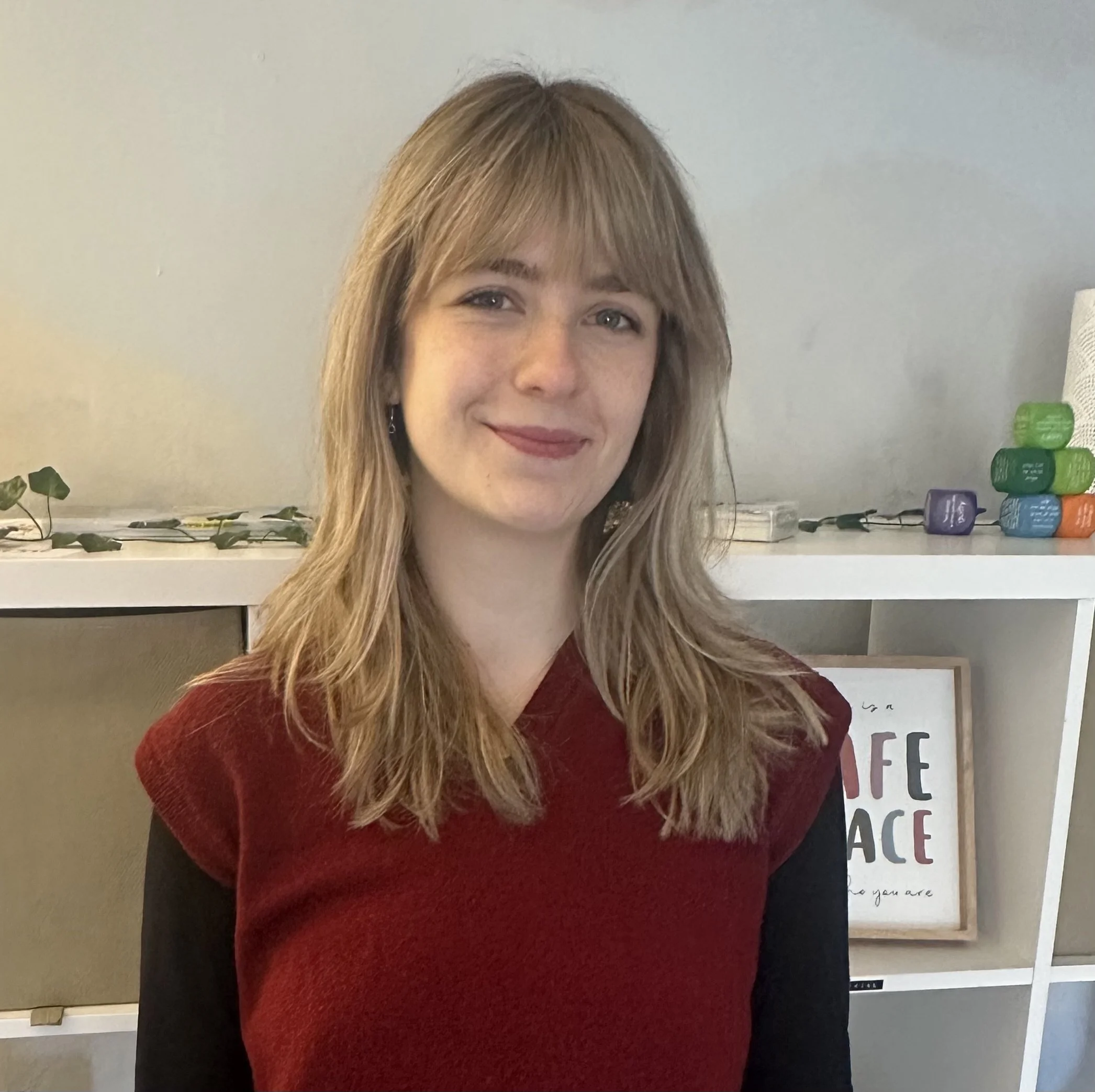 A young woman with shoulder-length blonde hair, wearing a red and black top, smiling in front of a white bookshelf with decorative items and framed art.