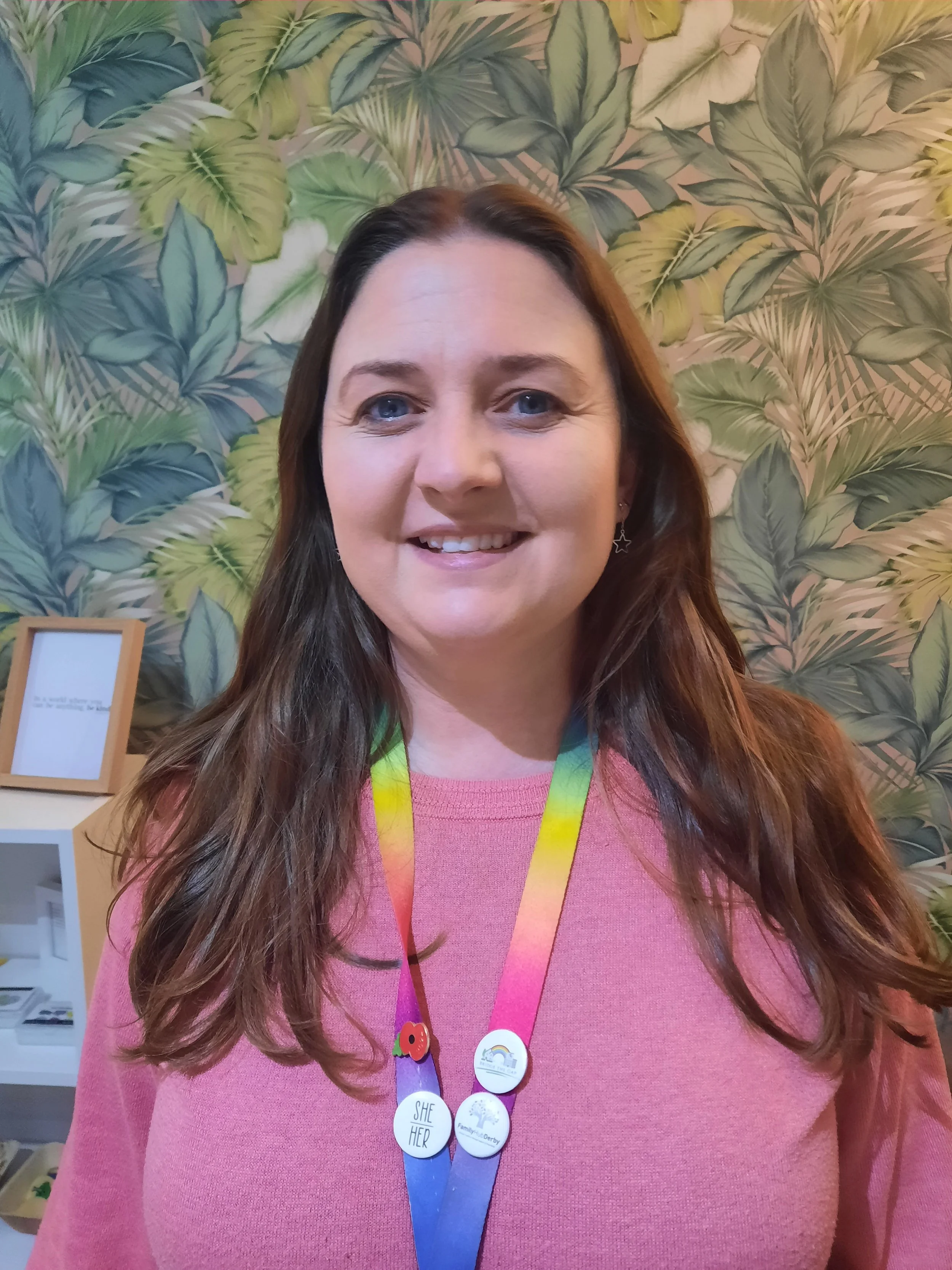 A woman with long brown hair, wearing a pink top, smiling, with a colorful lanyard with pins around her neck, standing in front of a leafy green and pink floral patterned wall.