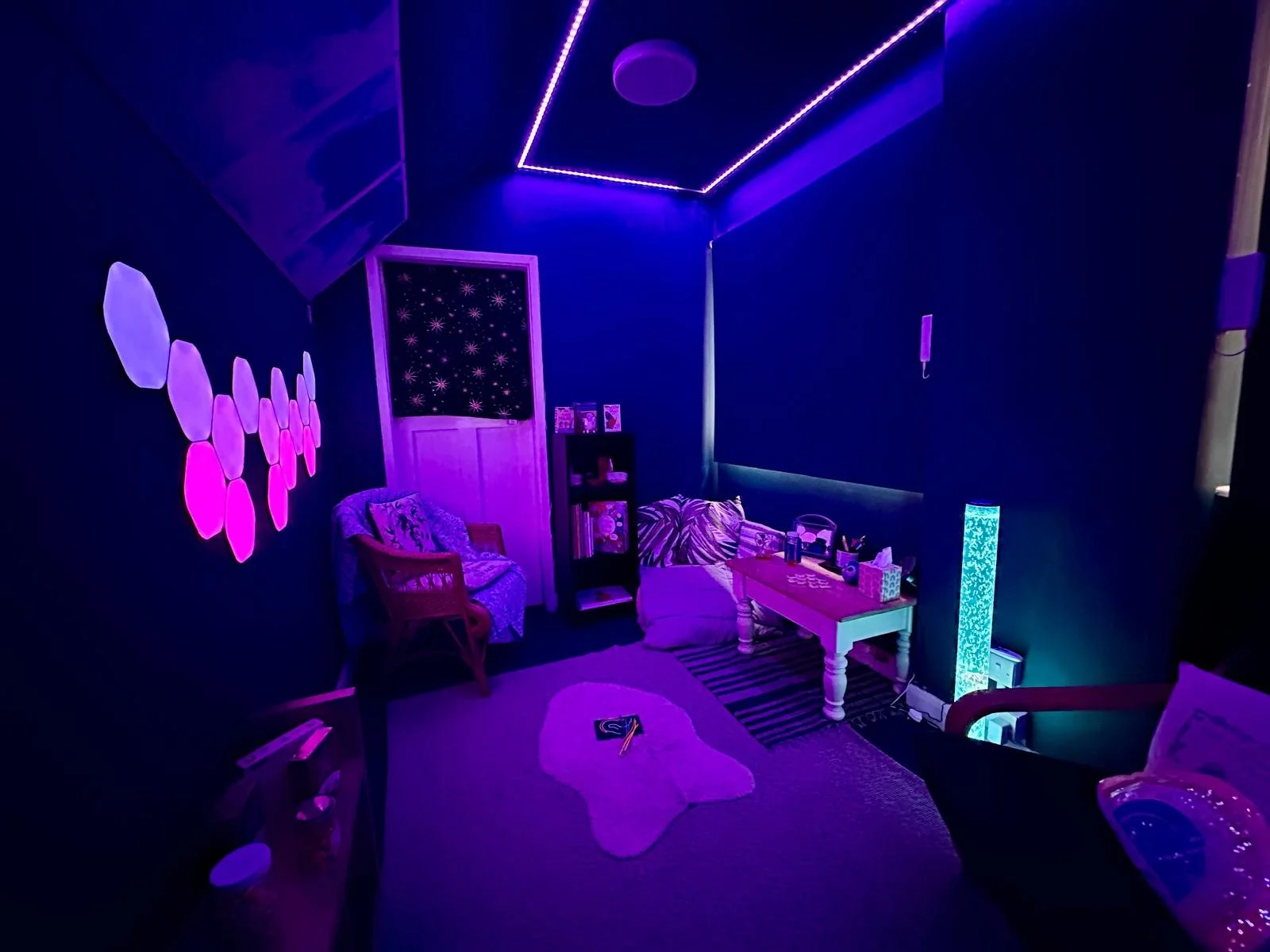 A dimly lit room with colorful neon and LED light decorations, a couch with patterned pillows, a small white table, a bookshelf, and wall art. The room has purple and blue lighting with various decorative items.